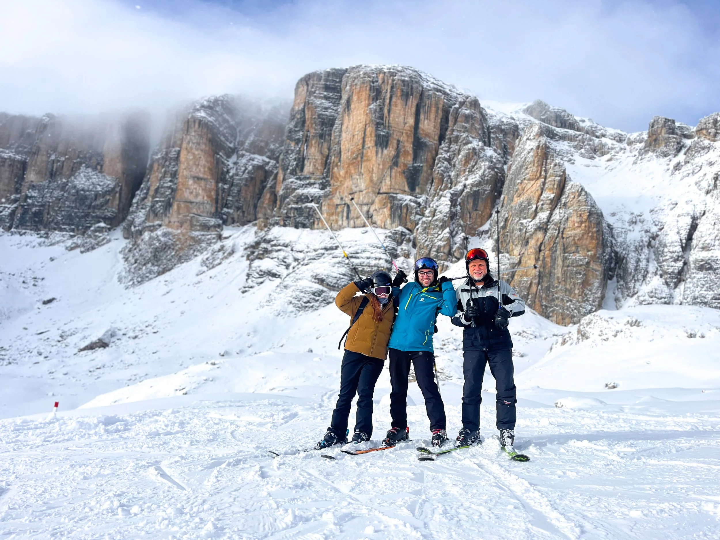 Three people standing on snow-covered ground with mountain cliffs in the background, smiling, holding ski poles and wearing ski gear and helmets.