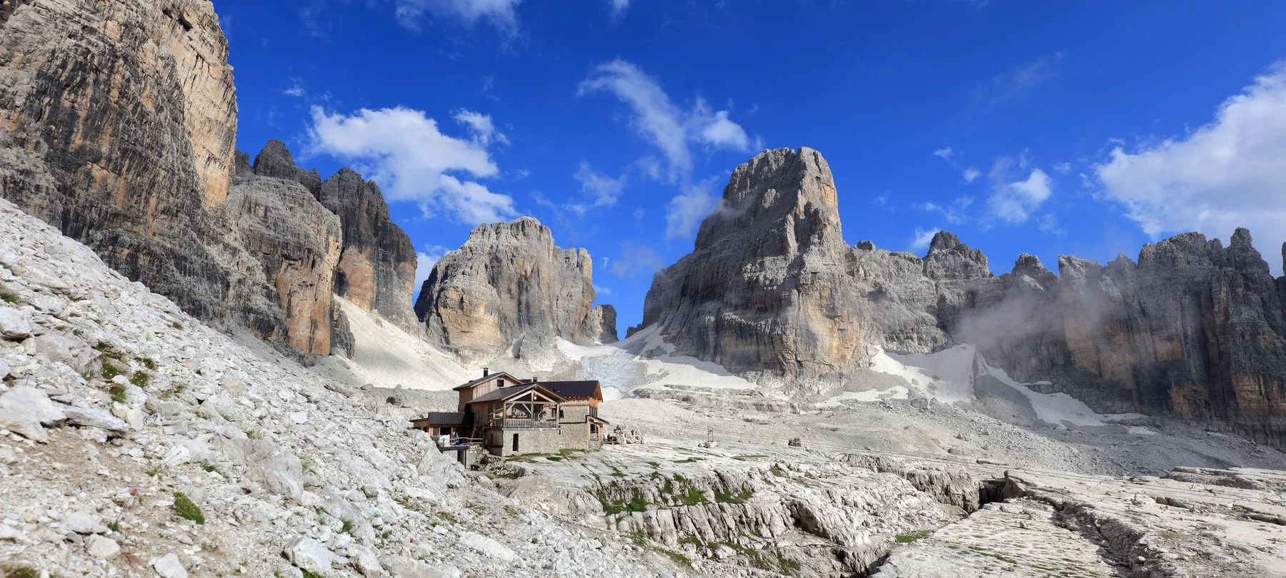 A mountain landscape with towering rocky peaks, a mountain hut in the foreground, and a bright blue sky with scattered clouds.