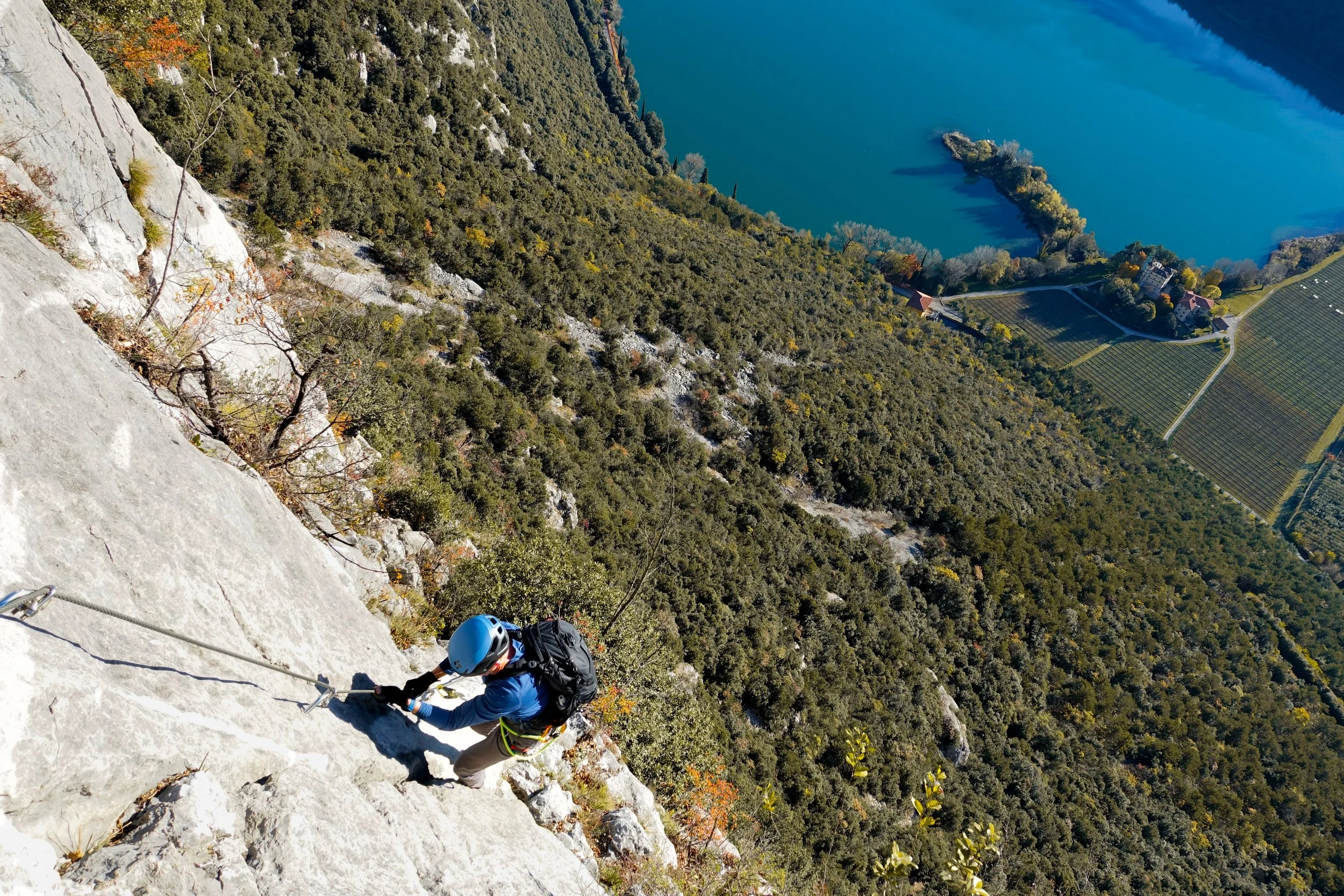 A person rock climbing on a steep mountain face, wearing a helmet and harness, with a lake, forest, and farmland in the background.