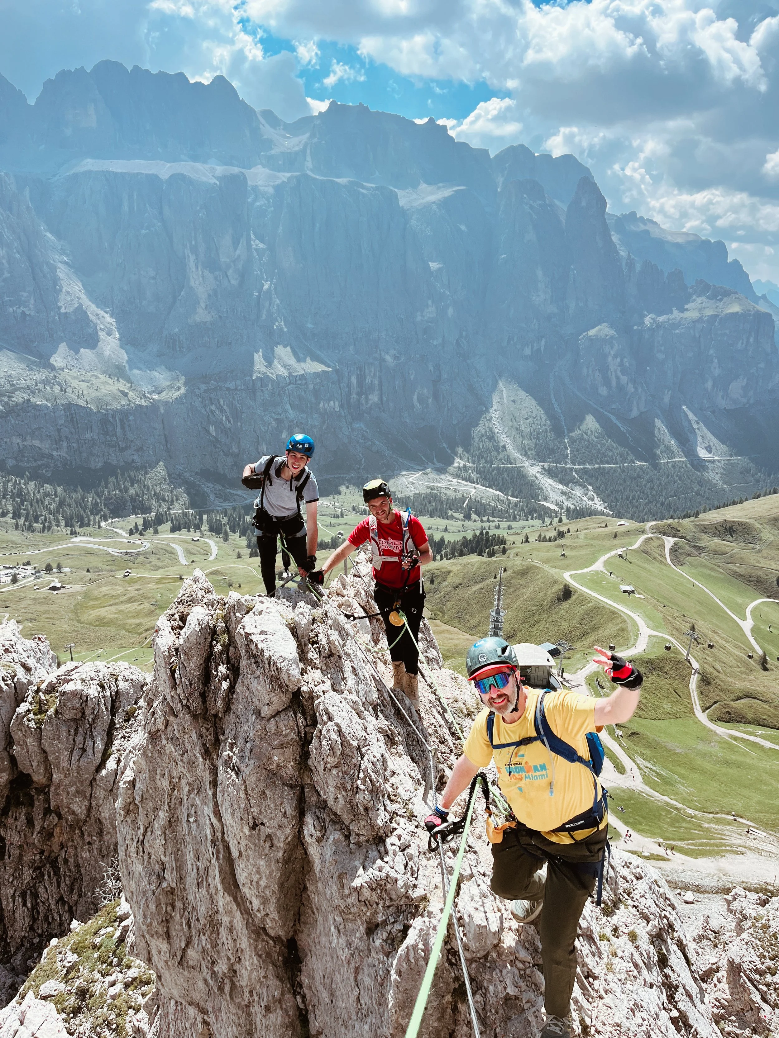 Three climbers wearing helmets and climbing gear on a rocky mountain ridge with a scenic view of mountains, green valleys, and winding roads below.