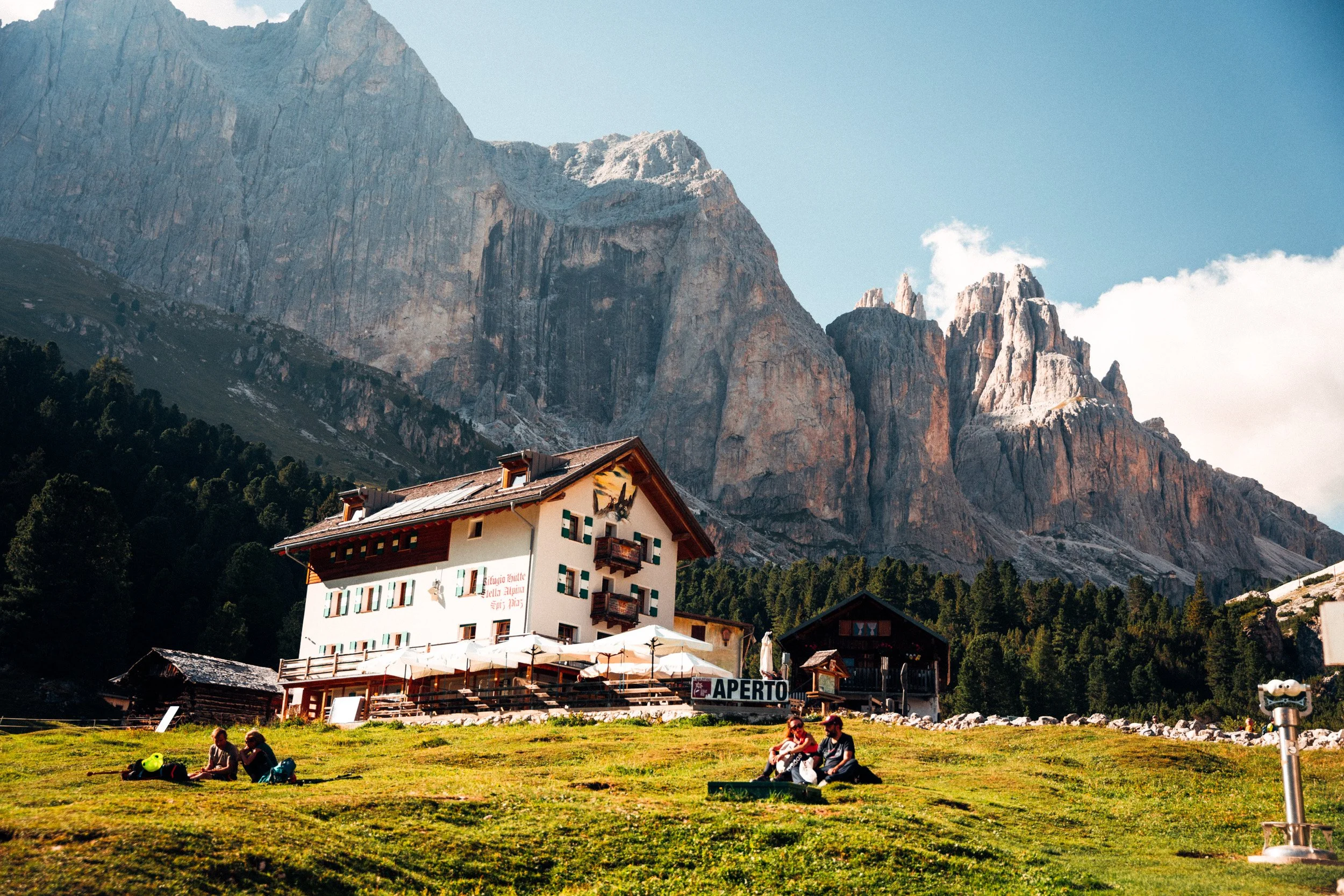 A mountain chalet with a large sign that reads 'APERT0', surrounded by green grass and trees, with towering mountains in the background. People are sitting on the grass, some with backpacks and others chatting, enjoying a sunny day.
