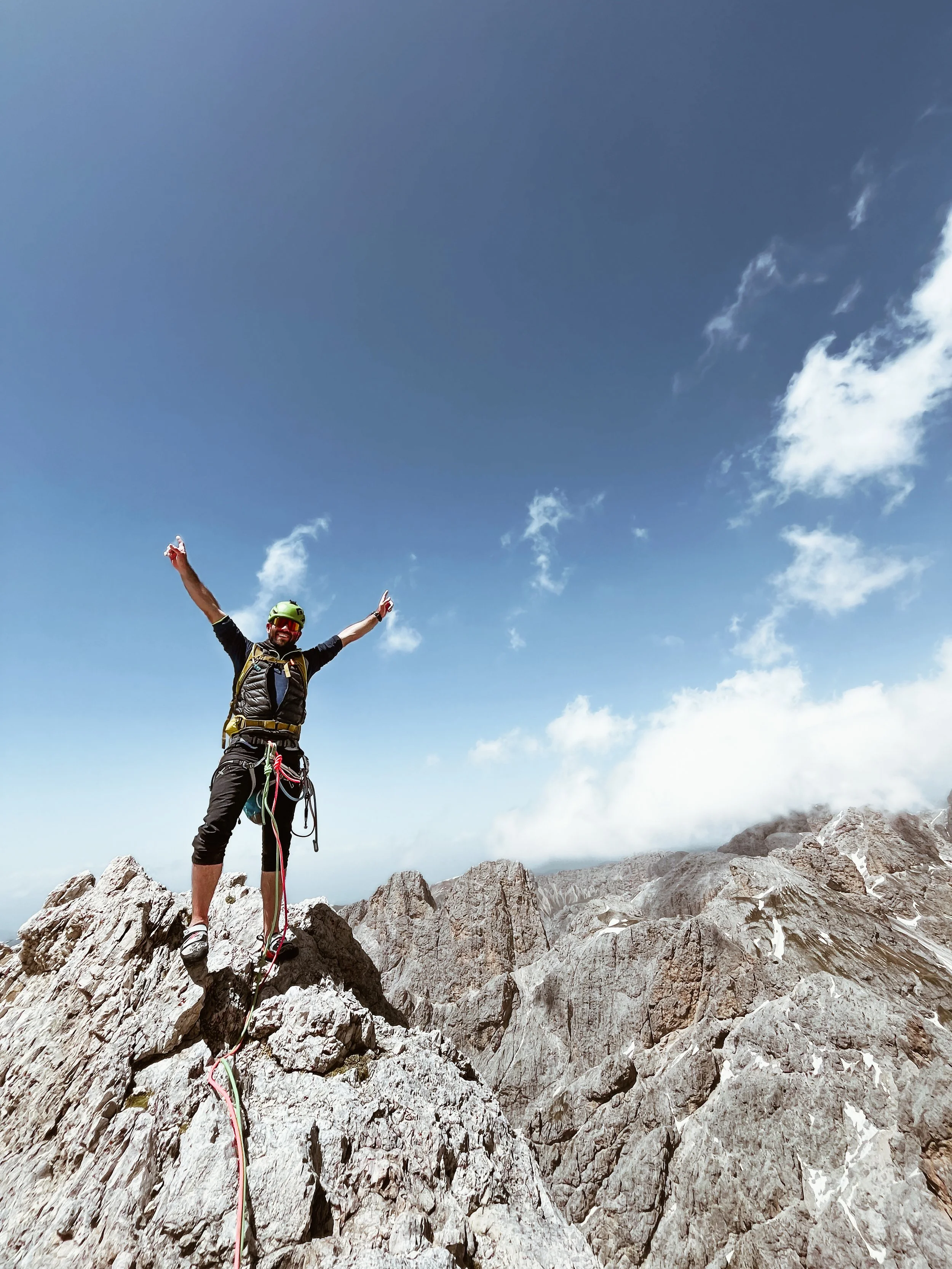 A man standing on top of a rocky mountain summit, raising his arms in victory, with a blue sky and clouds in the background.