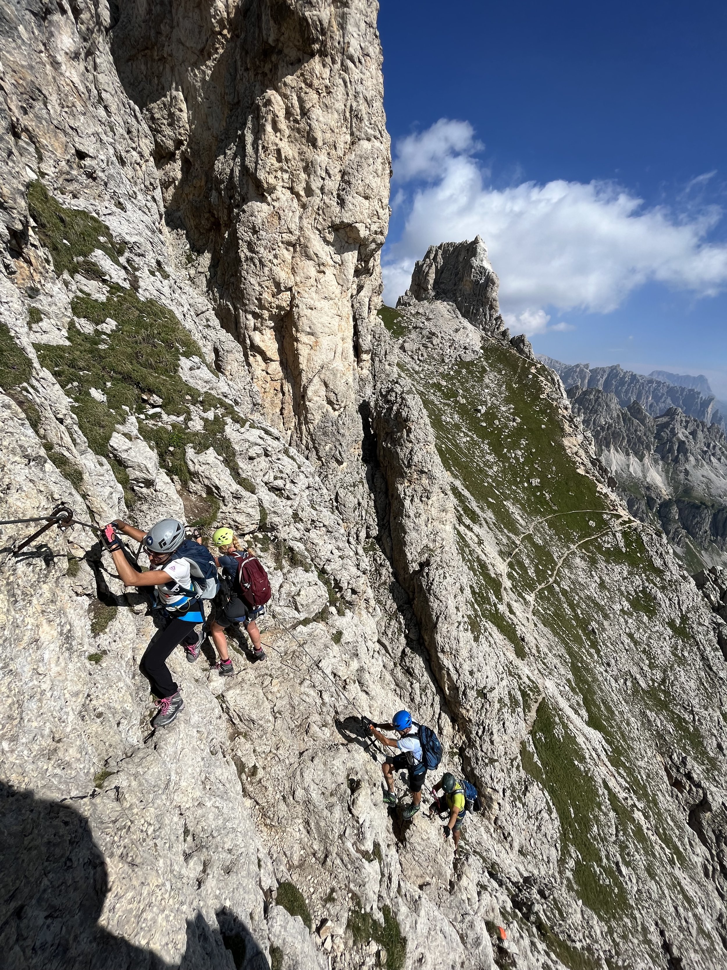 Mountaineers climbing a steep rocky mountain trail with safety ropes and helmets. They are climbing via ferrata Masarè following their guide Filippo Barbieri in the Rosengarten traverse dolomites safari.