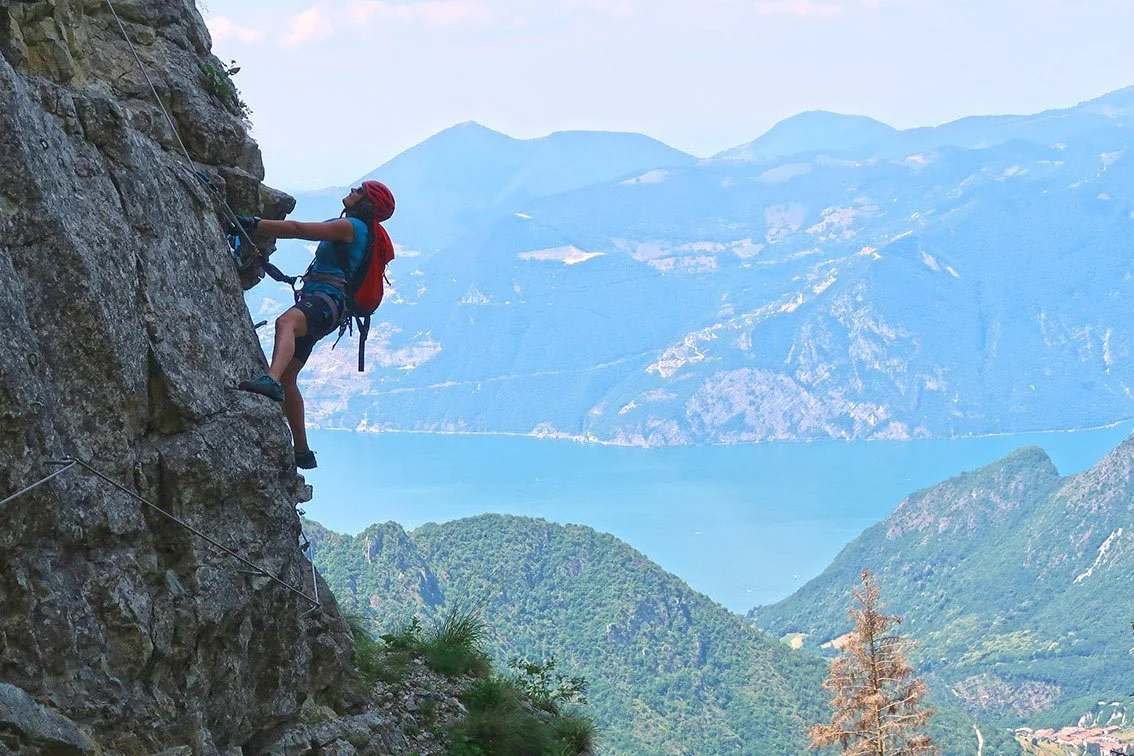 A person rock climbing on a steep cliff with a scenic lake and mountains in the background.