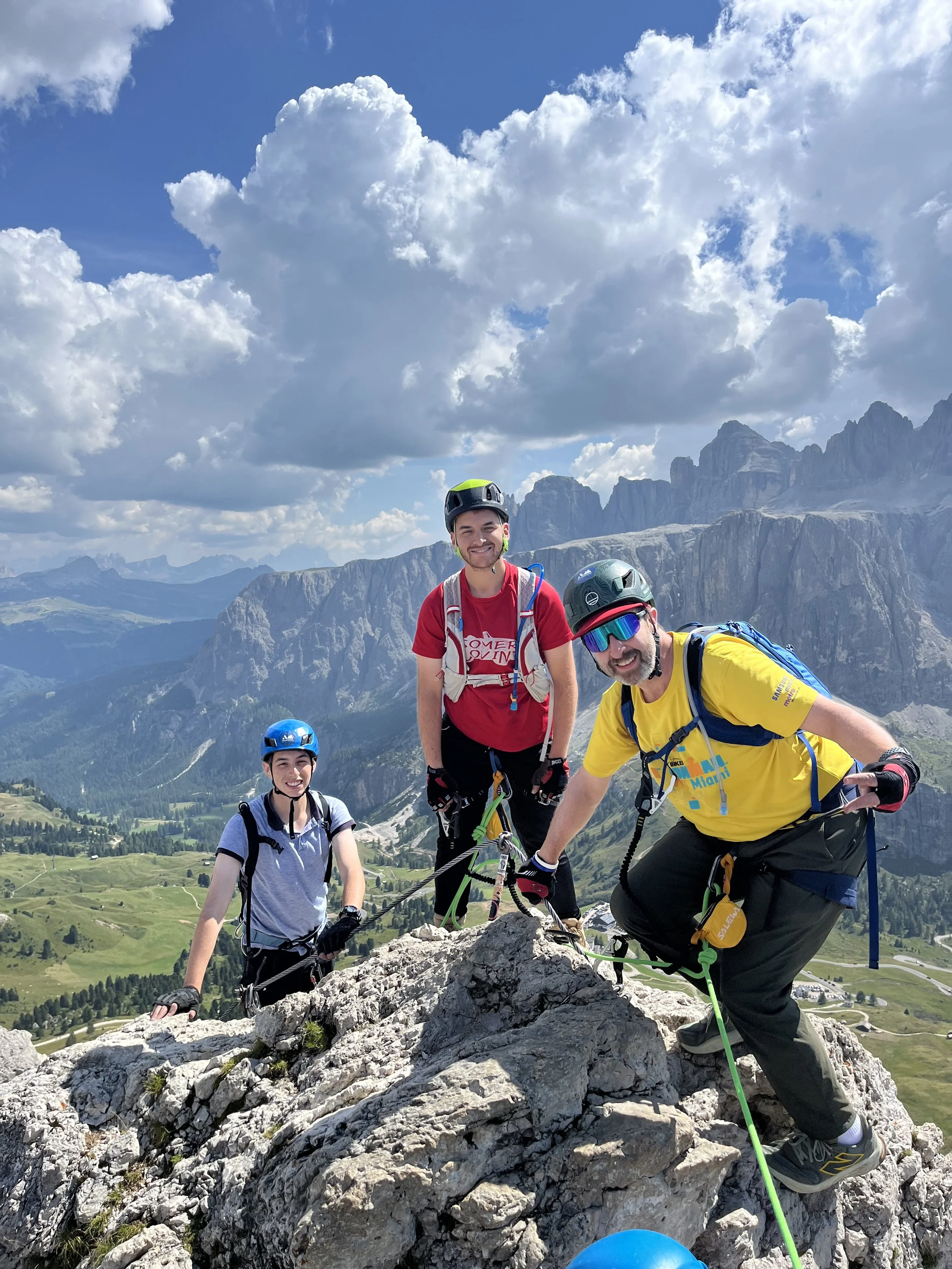 Three climbers wearing helmets and climbing gear on a rocky mountain ridge with a scenic view of mountains, green valleys, and winding roads arriving at Pass Gardena. They are climbing Piccolo Cir, in the Dolomites following their guide Filippo