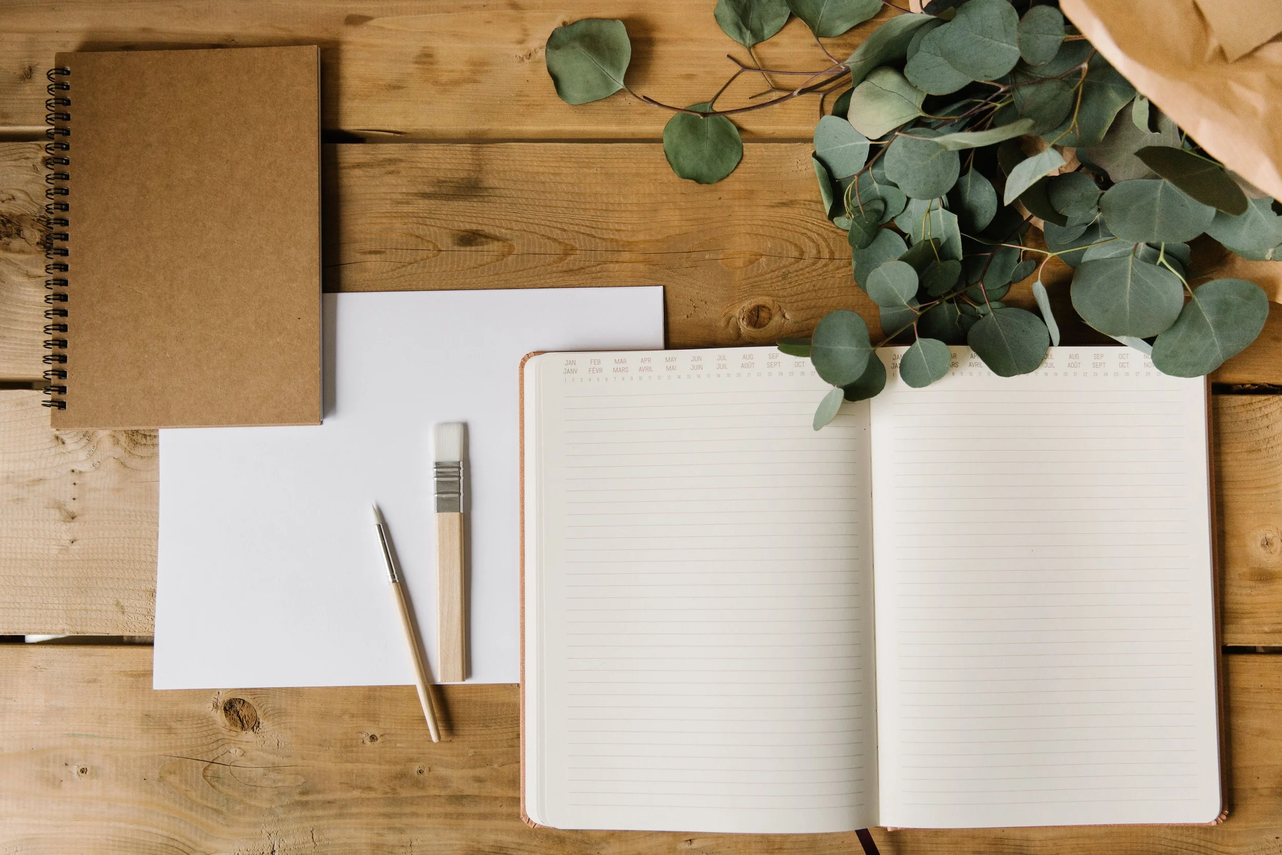 Un carnet en papier kraft, un agenda avec des pages blanches, un pinceau, un petit sac en papier avec des feuilles vertes, un calendrier ou une fiche de planification, le tout disposé sur une surface en bois.