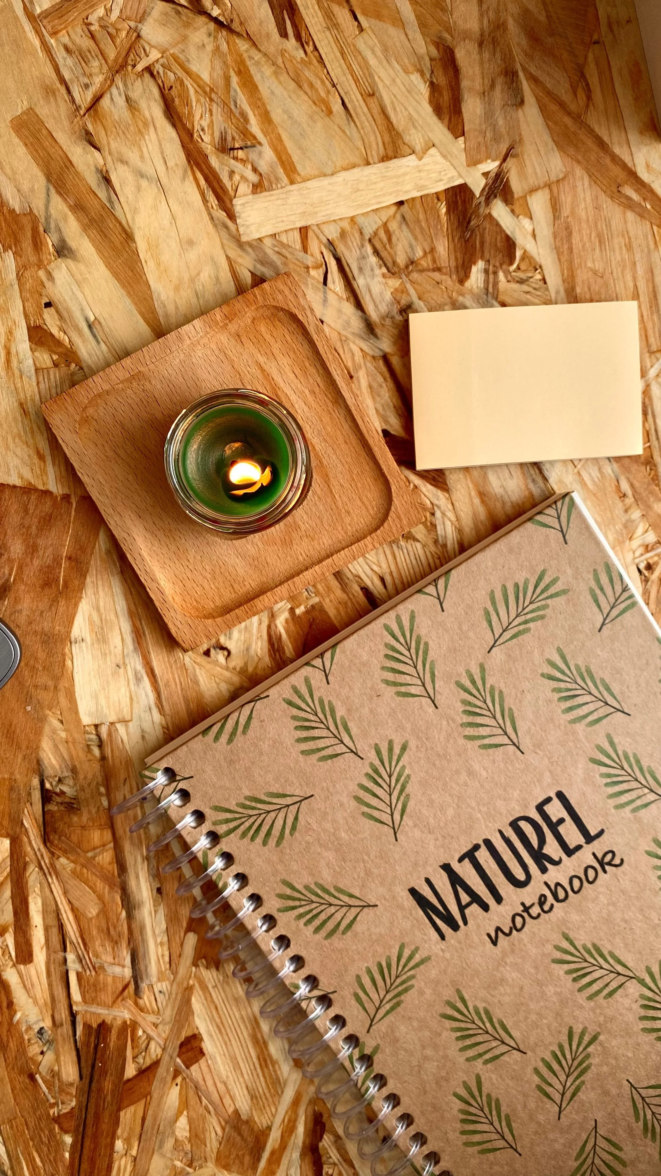 Une photo d'un carnet de notes à couverture marron avec des branches vertes, un carnet à spirale, une enveloppe, une bougie allumée dans un verre, et quelques feuilles de papier sur un plan de travail en bois.