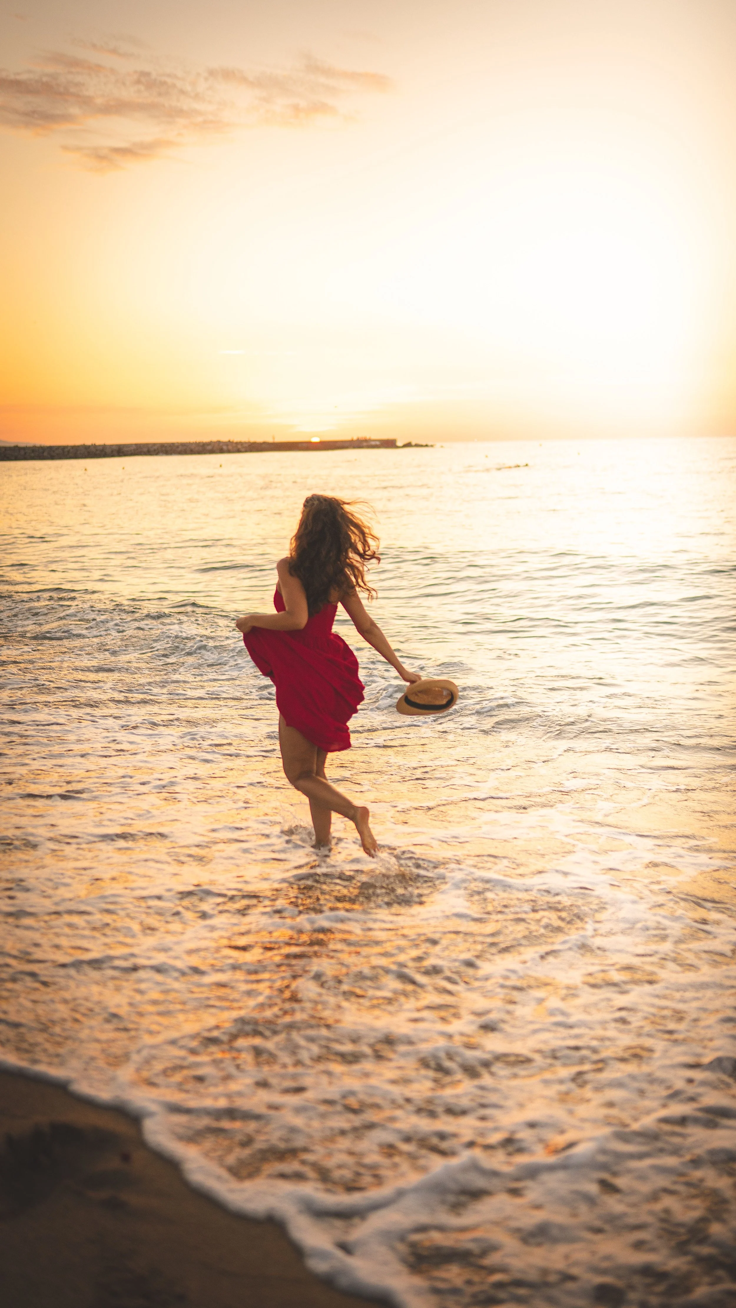 Une femme en robe rouge joue dans l'eau de la mer au coucher du soleil, tenant un chapeau en main, à la plage.