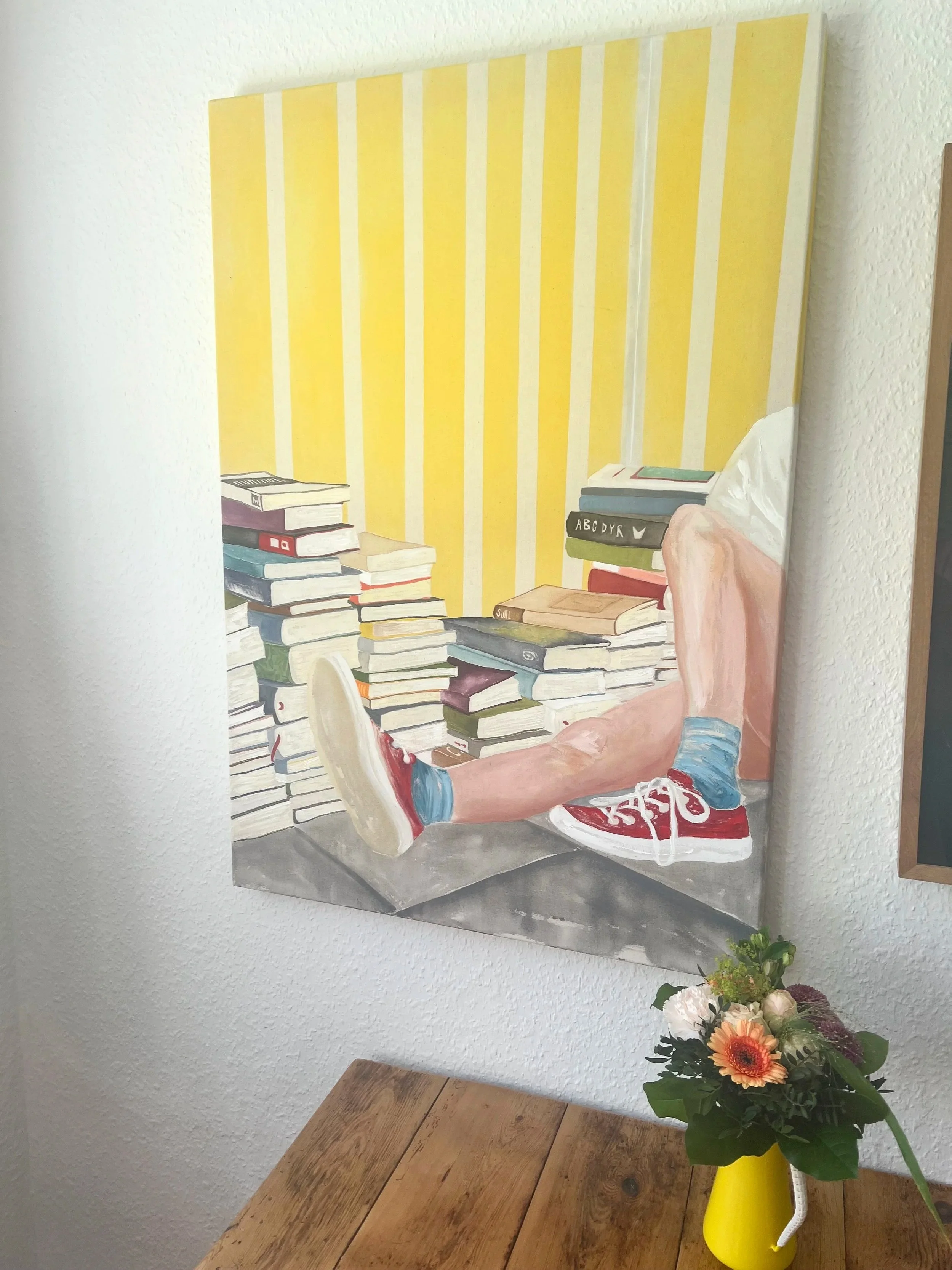 A painting of a person sitting on the floor with their legs stretched out, surrounded by piles of books. The person's face is not visible, and they are wearing red sneakers, blue socks, and shorts. The background features yellow and white vertical st