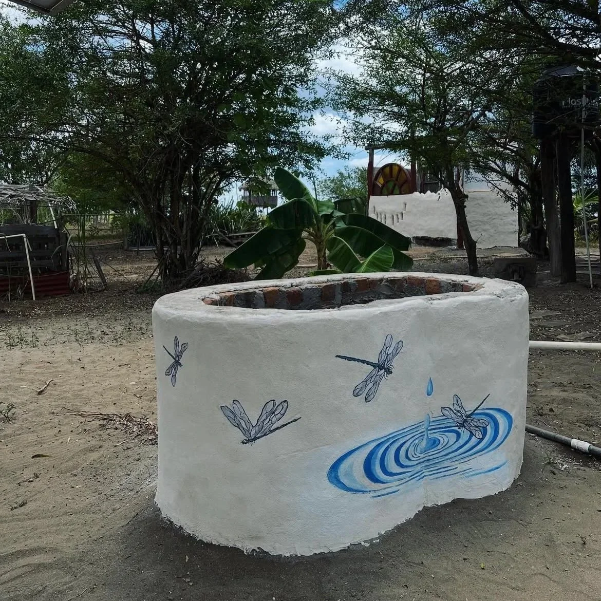 Painted white outdoor fountain with dragonflies and water ripples design, surrounded by trees and plants