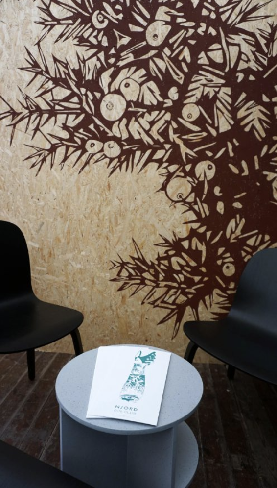 An interior space with a decorative wall featuring a large, abstract plant or tree design in dark brown against a light background. Two black chairs are positioned on either side of a small round table with a brochure on top.