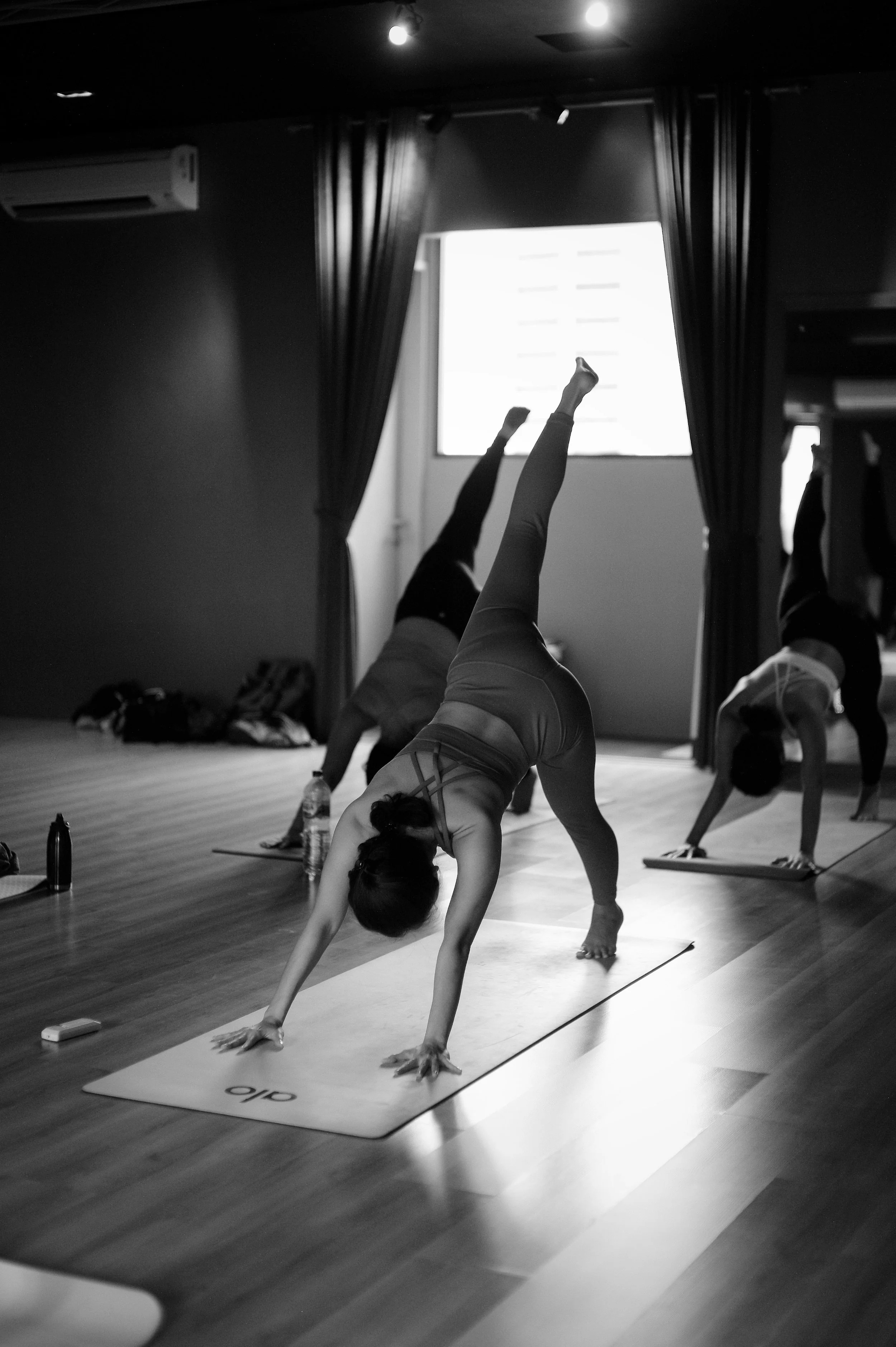 A group of people practicing yoga indoors, with one person in the foreground performing a handstand variation on a yoga mat, and others in the background doing different poses.