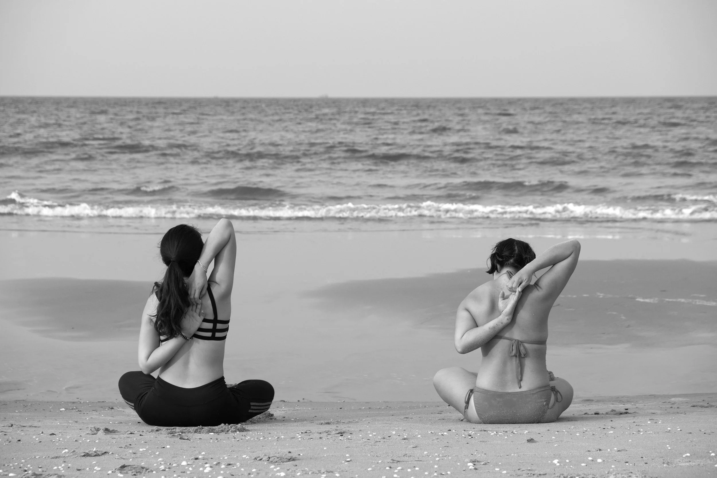 Two women practicing yoga on the beach, sitting cross-legged facing the ocean, with one arm bent behind their head and the other reaching over their shoulder, in black and white.