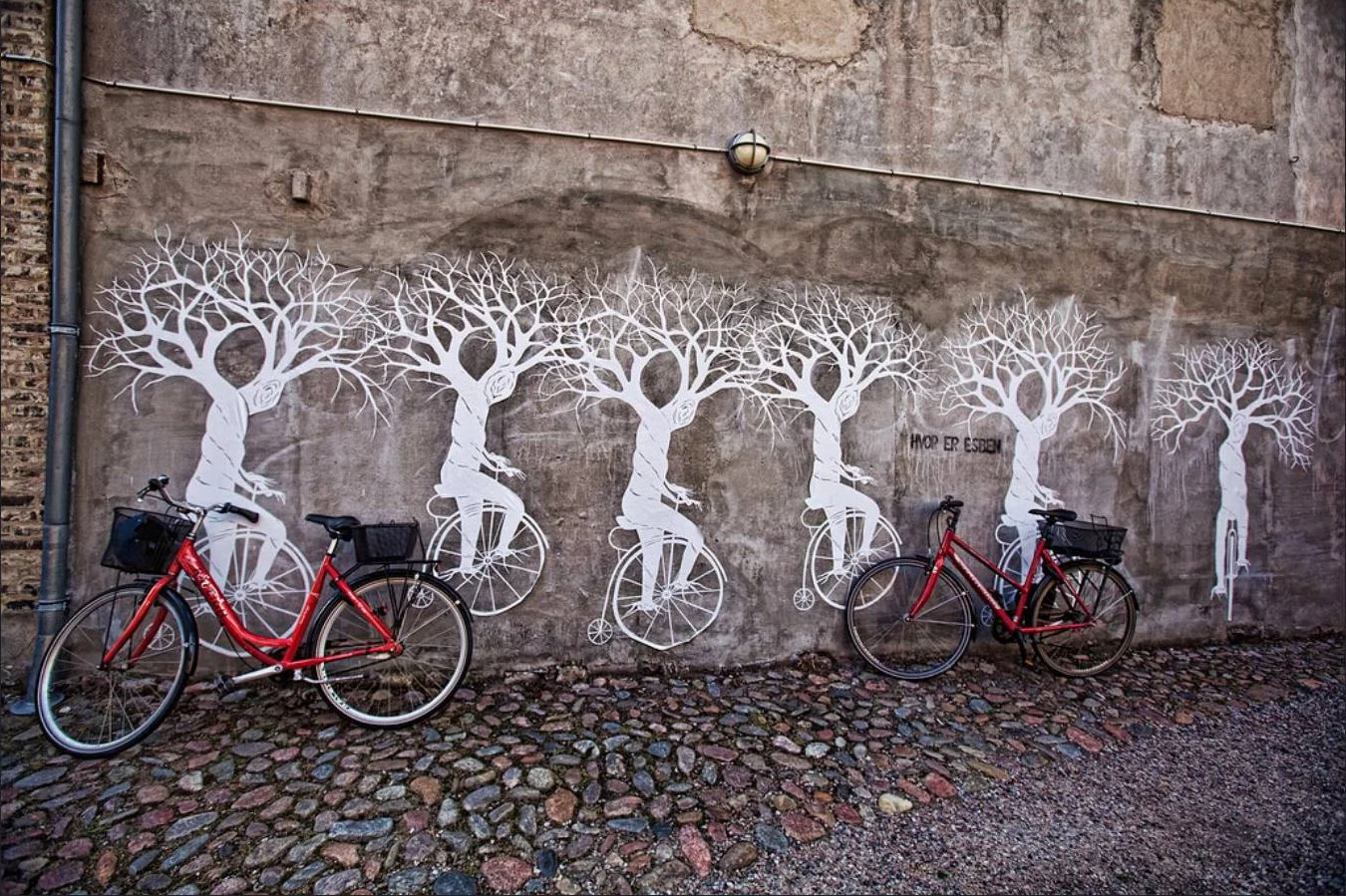 Street art mural of white trees with bicycle wheels and figures on a textured concrete wall, with two red bicycles parked in front.