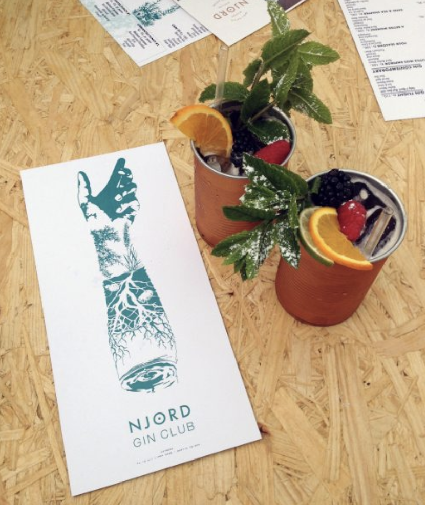Two cocktails garnished with fruit and mint, placed on a wooden table next to a white paper menu from NJORD Gin Club and some receipts.