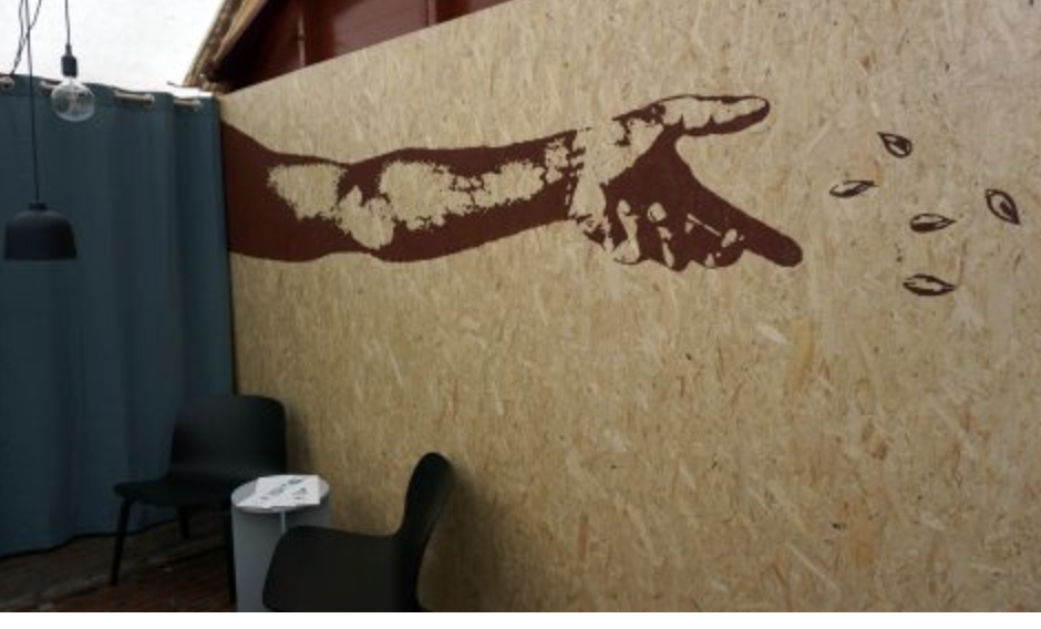 Wooden wall with a mural of a pointing hand and some small leaf-like objects, two black chairs, a white side table, and blue curtains.