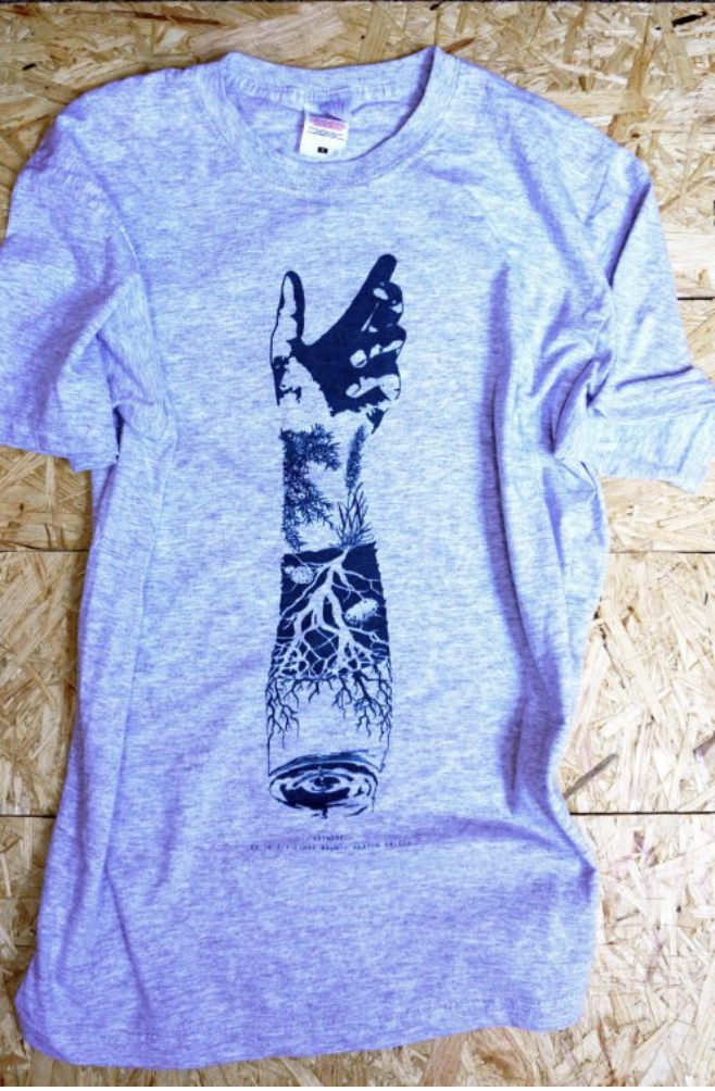 Gray T-shirt with black graphic of a hand pointing upward, containing tree, roots, and eye illustrations, laid on wood background.