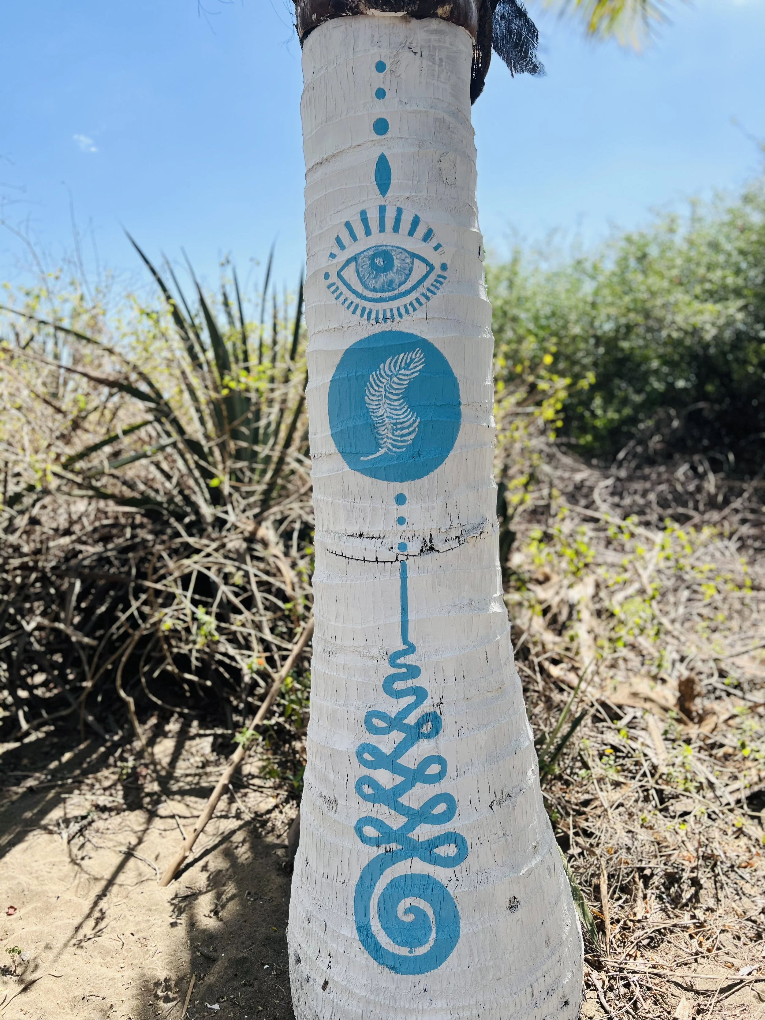 A painted white palm tree trunk with blue symbols, including an eye, a fern leaf, and decorative swirl and line designs, outdoors with bushes and a blue sky in the background.