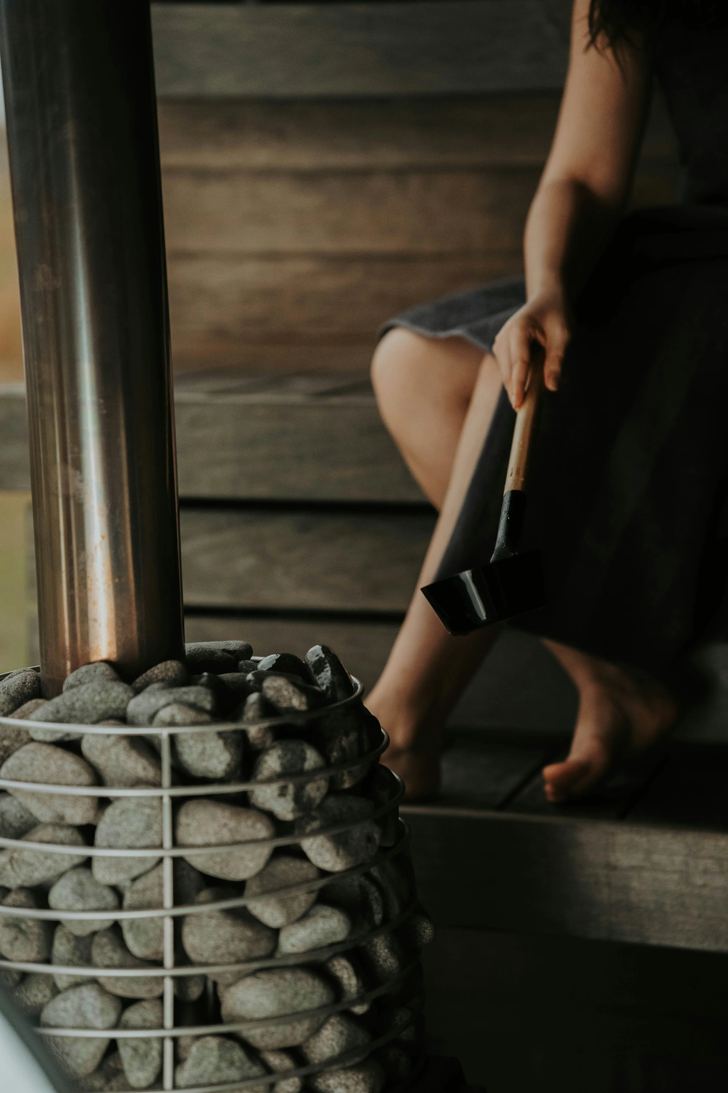 Person sitting near a sauna heater with rocks, holding a ladle.