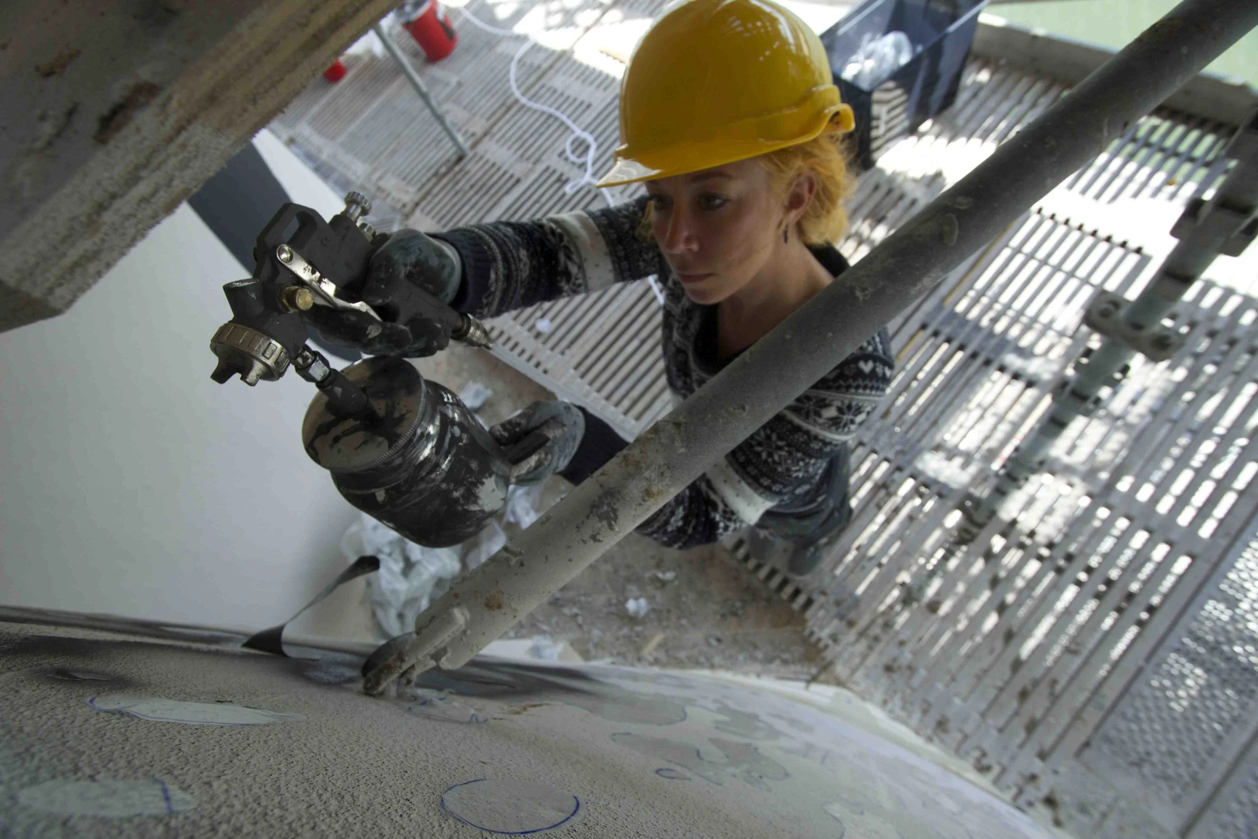 A worker wearing a yellow safety helmet and gloves is using a spray gun to apply paint on a wall from scaffolding.
