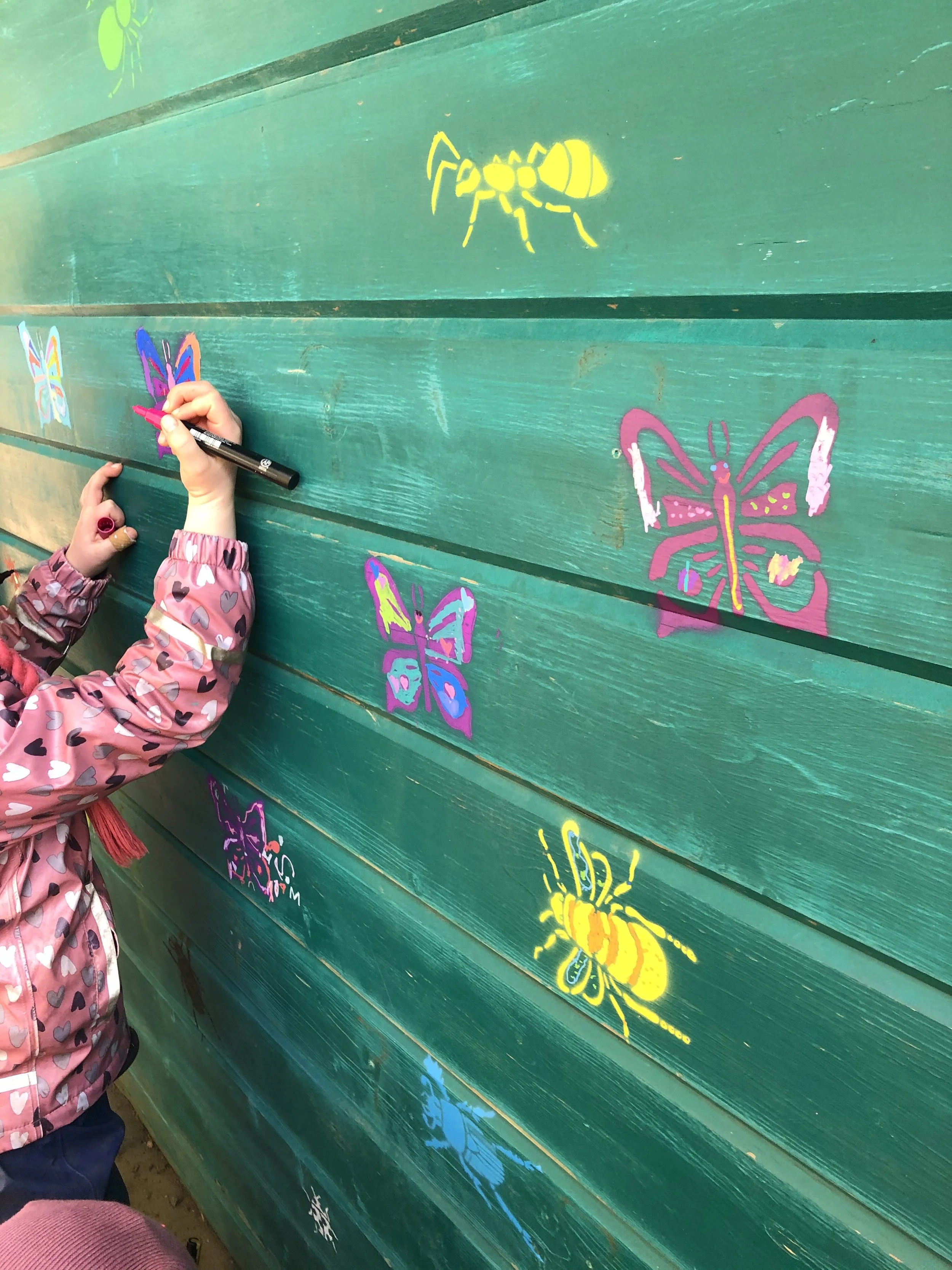 Child drawing colorful butterfly illustrations on a green wooden wall.