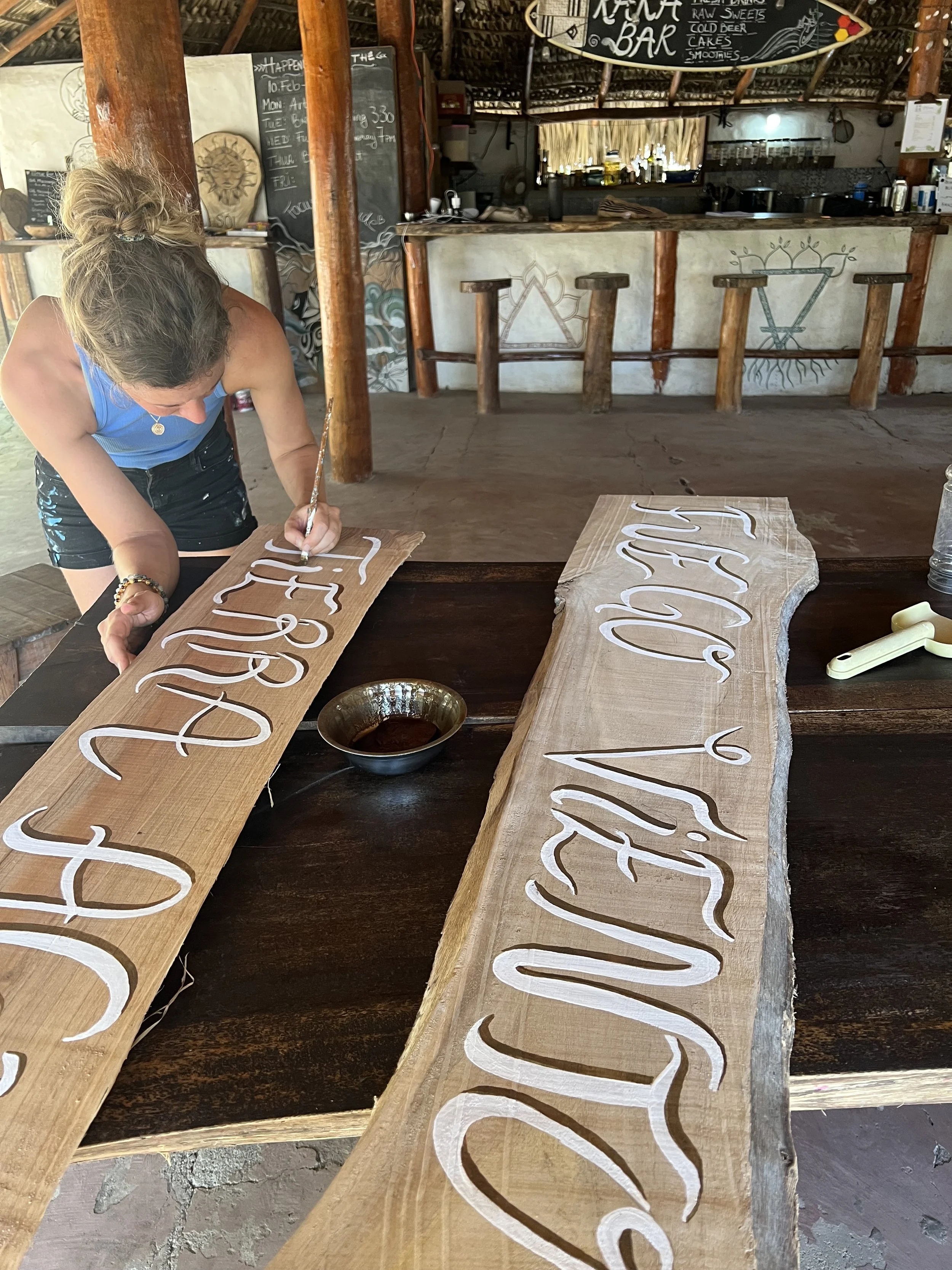 A woman is painting white lettering on two long wooden signs at a rustic bar or cafe. The signs appear to have handwritten script, and one reads 'Bar' while the other seems to start with 'Welcome' or similar. Behind her, there is a bar area with stoo