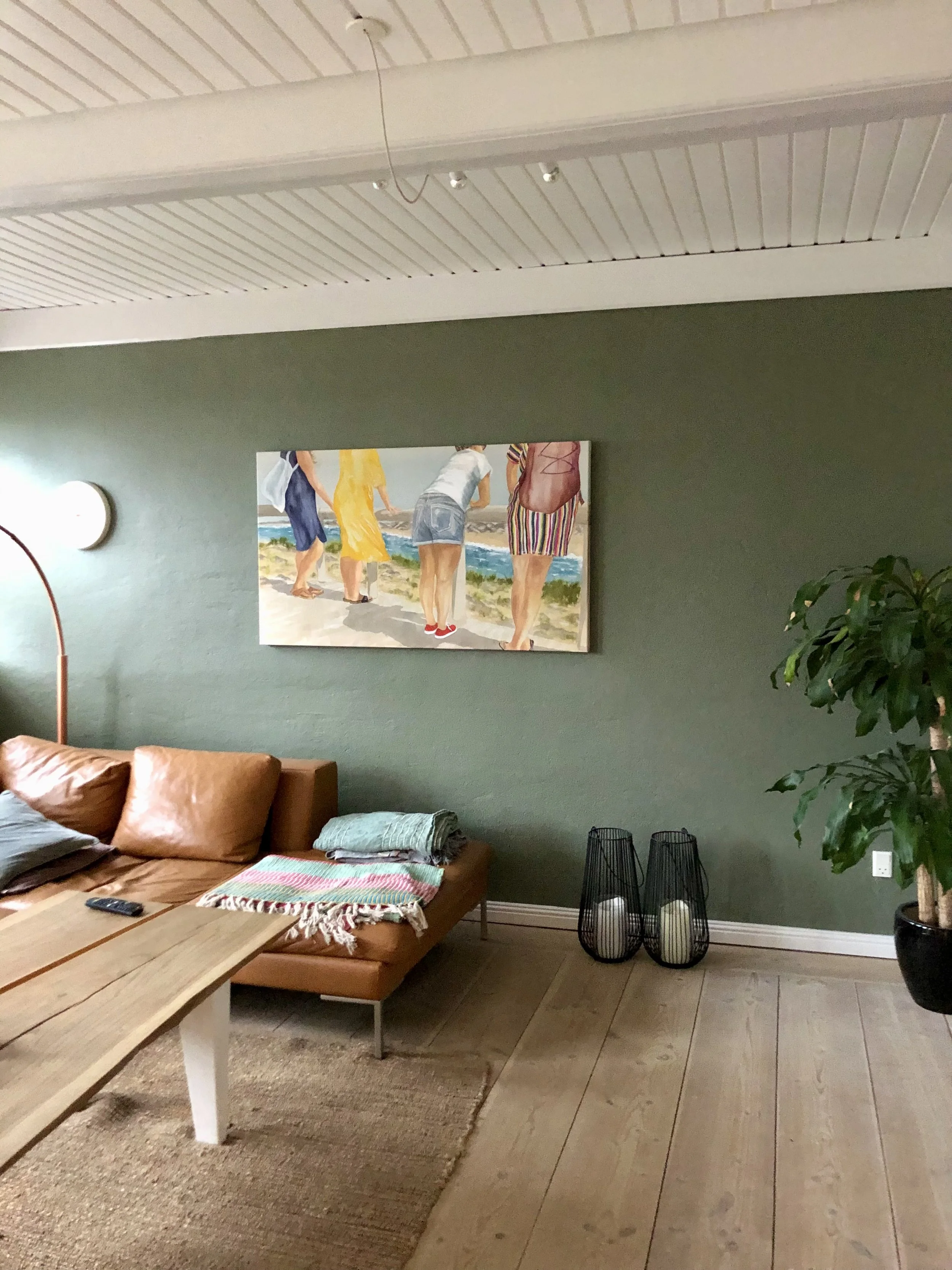 Living room with tan leather sofa, throw pillows, a wooden coffee table, decorative lamps, a large green potted plant, and a painting of people walking on the beach hanging on the green wall.