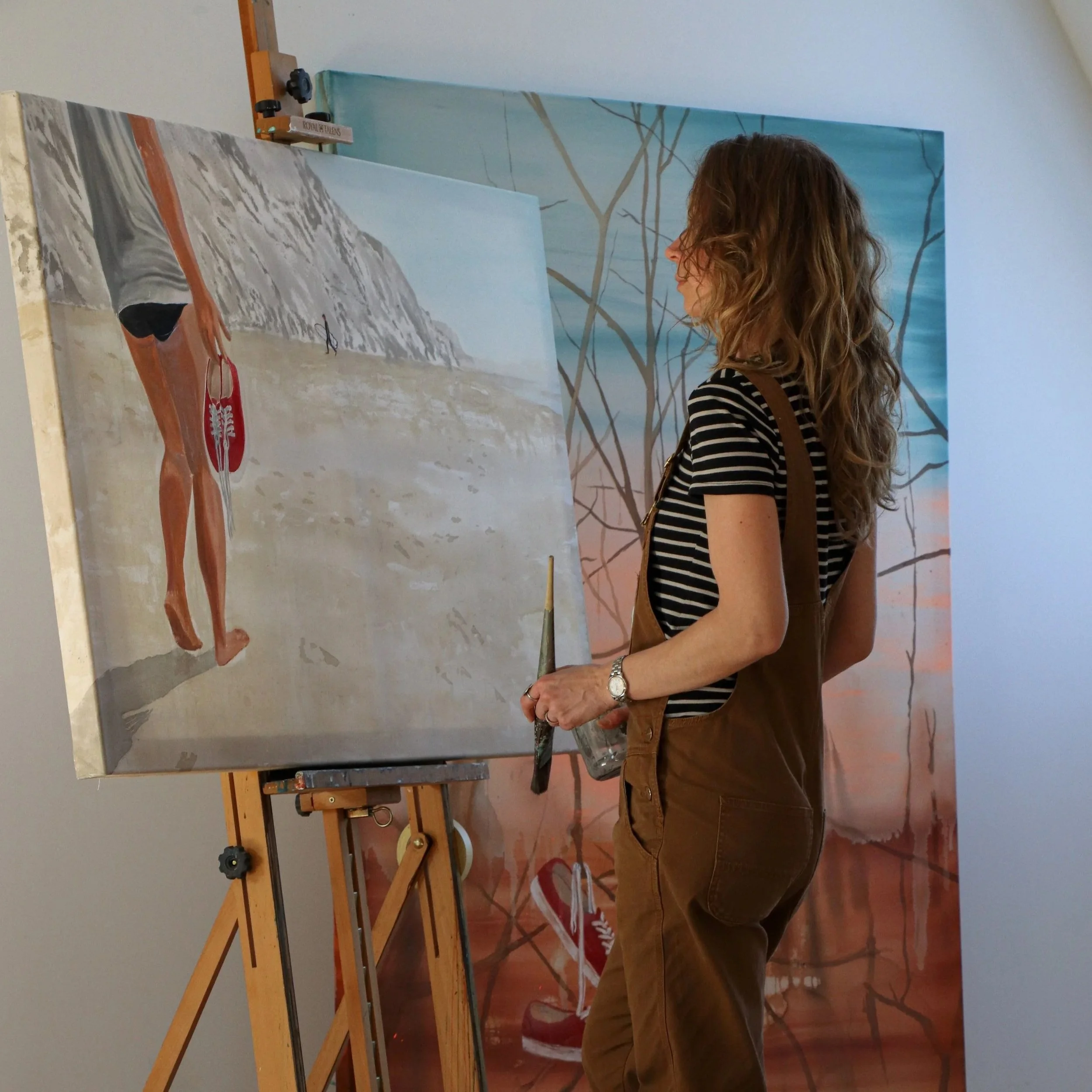 A woman with wavy brown hair wearing a black and white striped shirt and brown overalls, standing in front of easels and paintings, holding a paintbrush and glass jar, in an art studio.