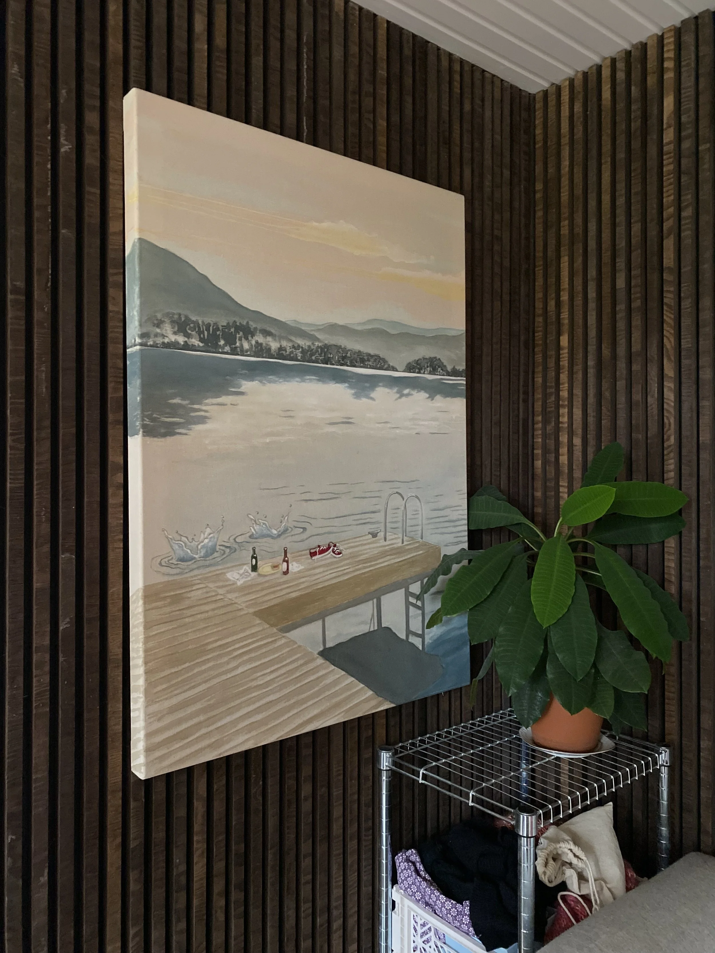 Painting of a lakeside scene with mountains in the background, featuring a dock with drinks and people jumping into the water, hanging on a dark wooden wall with a green potted plant on a metal shelf below.