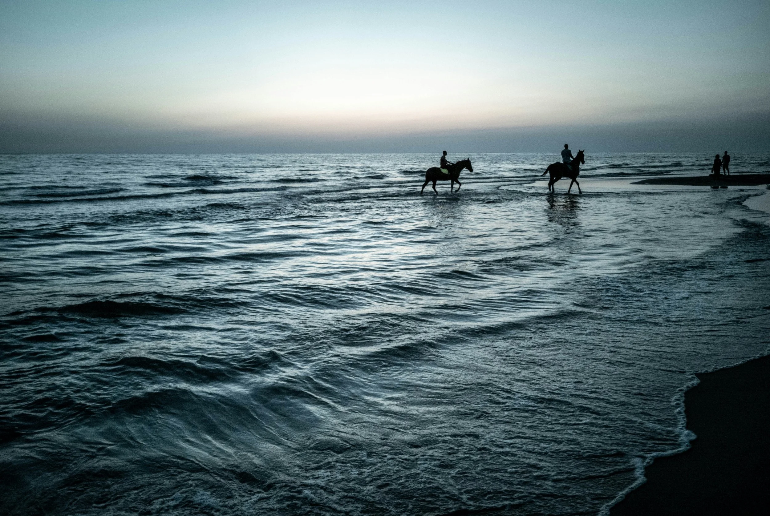 People riding horses on the beach at sunset.