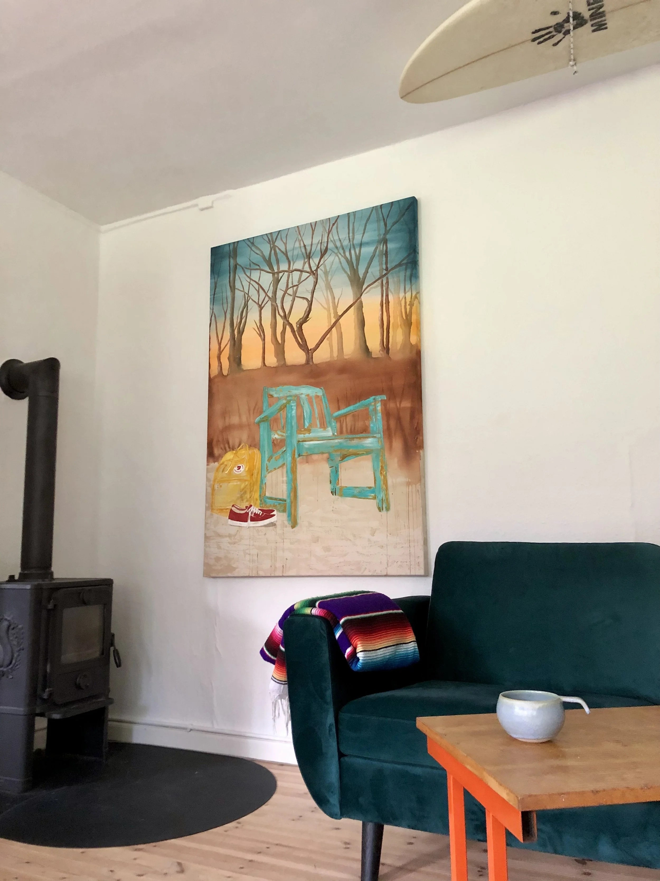 Living room with a green sofa, a colorful striped blanket, a wooden coffee table with a white bowl, a painting of a sunset with trees and a blue chair, a wood stove, and surfboards hanging from the ceiling.