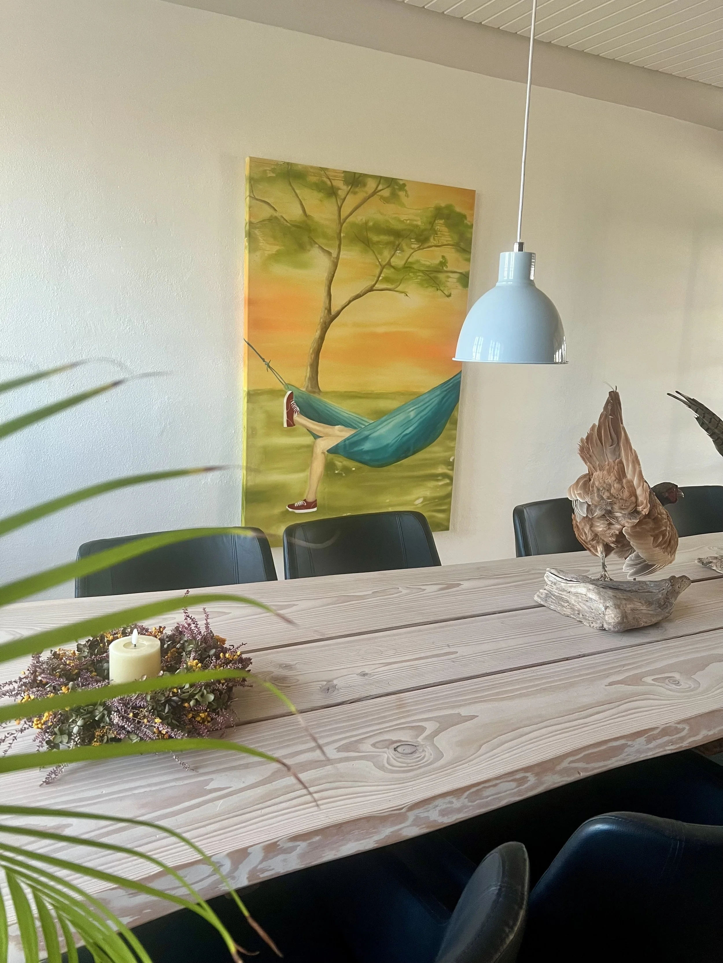 A wooden dining table with a lamp, a flower arrangement, and a hen sculpture on it. There is a painting of a person in red shoes in a hammock under a tree on the wall behind. The wall has a white textured surface.