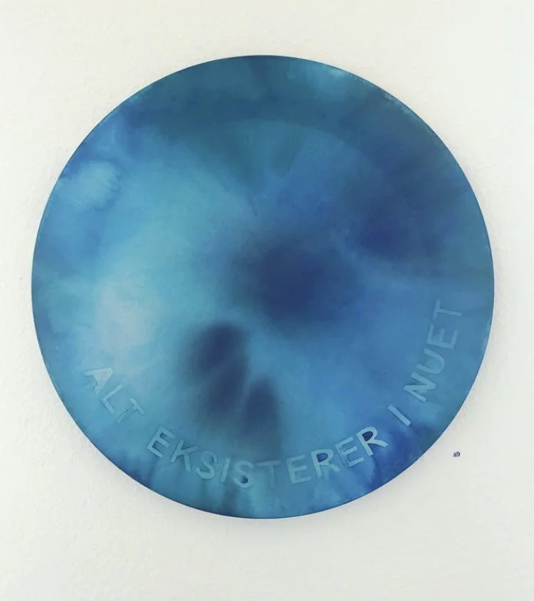 Blue circular artwork with the German phrase "Auf Existerer I Nacht" inscribed around it, displayed on a white wall.