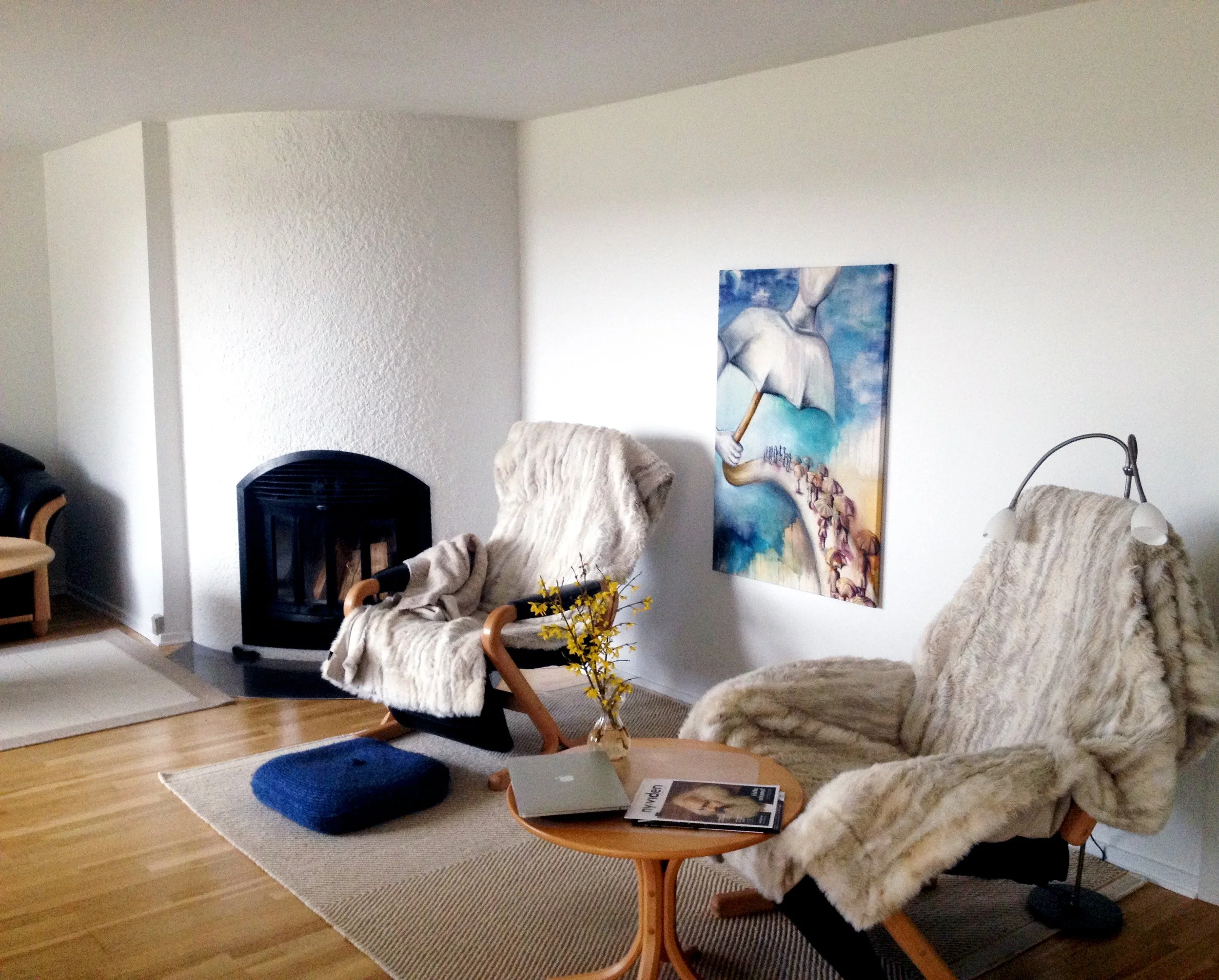 Living room with two beige fur-covered armchairs, a small wooden table with a magazine and laptop, a blue footstool, a painting of an umbrella, and a fireplace.