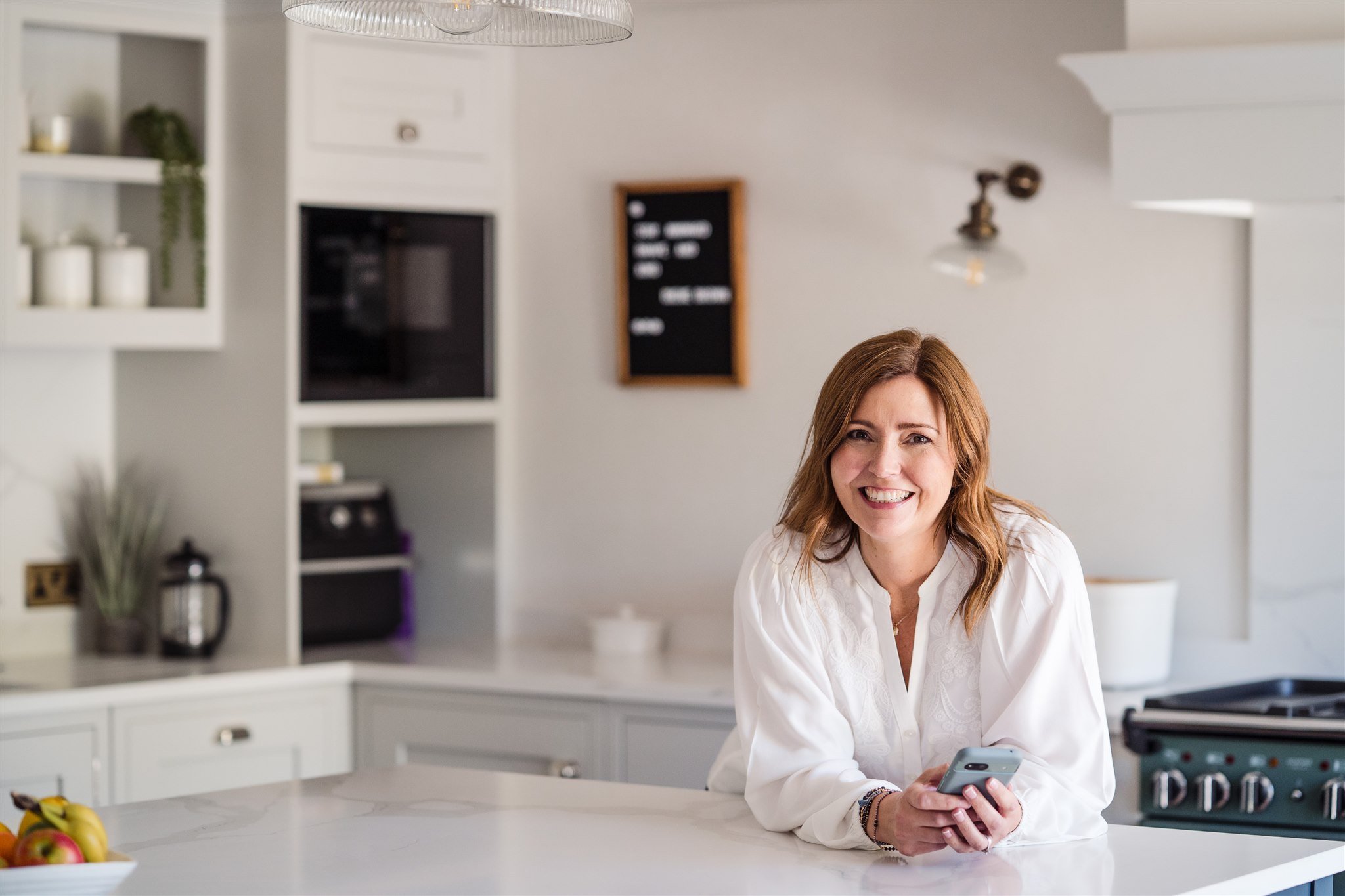 Female leadership and neurodiversity coach with brown hair smiling and holding a smartphone in a modern kitchen with soft grey cabinets and a marble countertop.