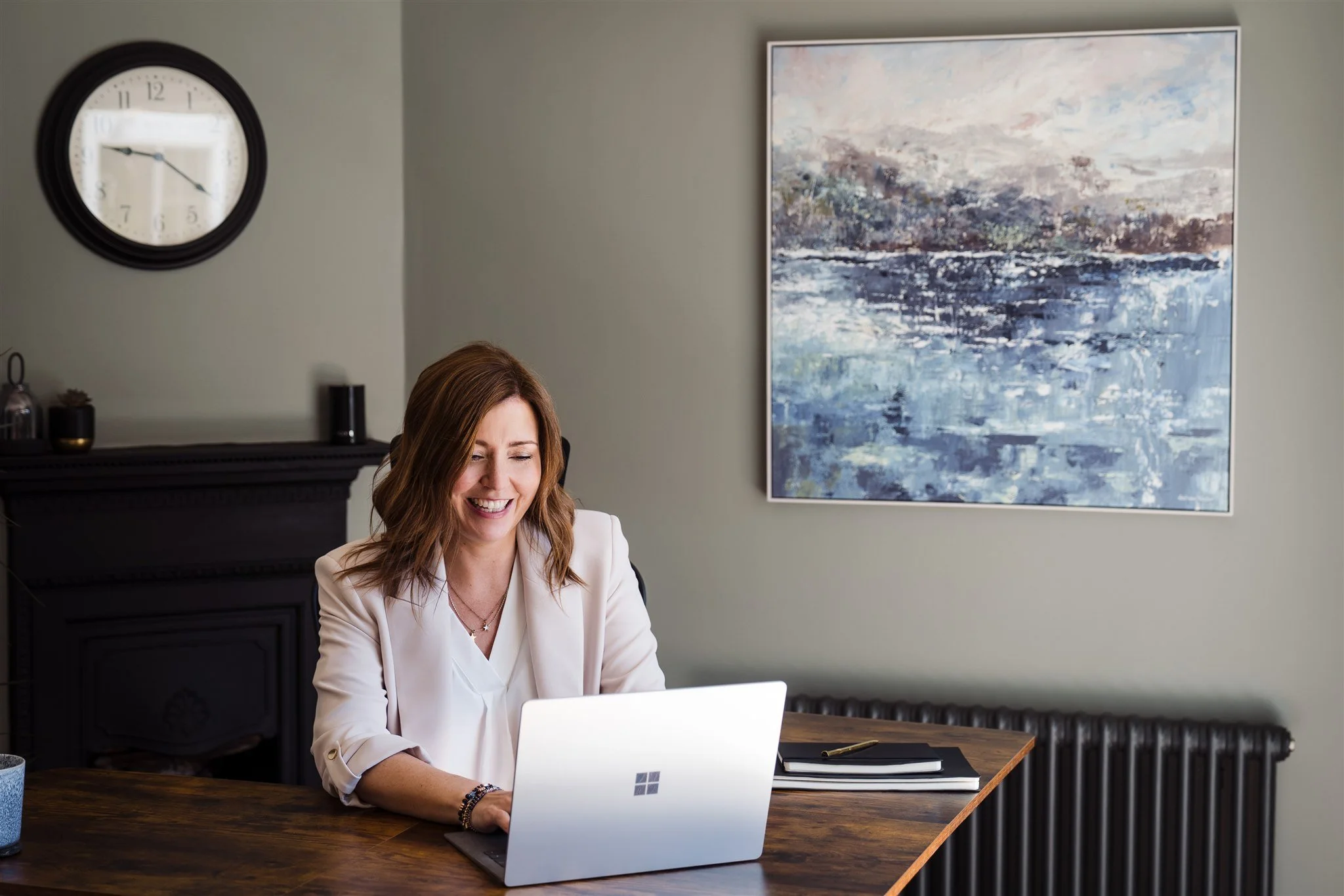 A female leadership and neurodiversity coach with shoulder-length brown hair sat at a wooden desk. She is smiling, looking like she is in coversation with someone virtually on her laptop.