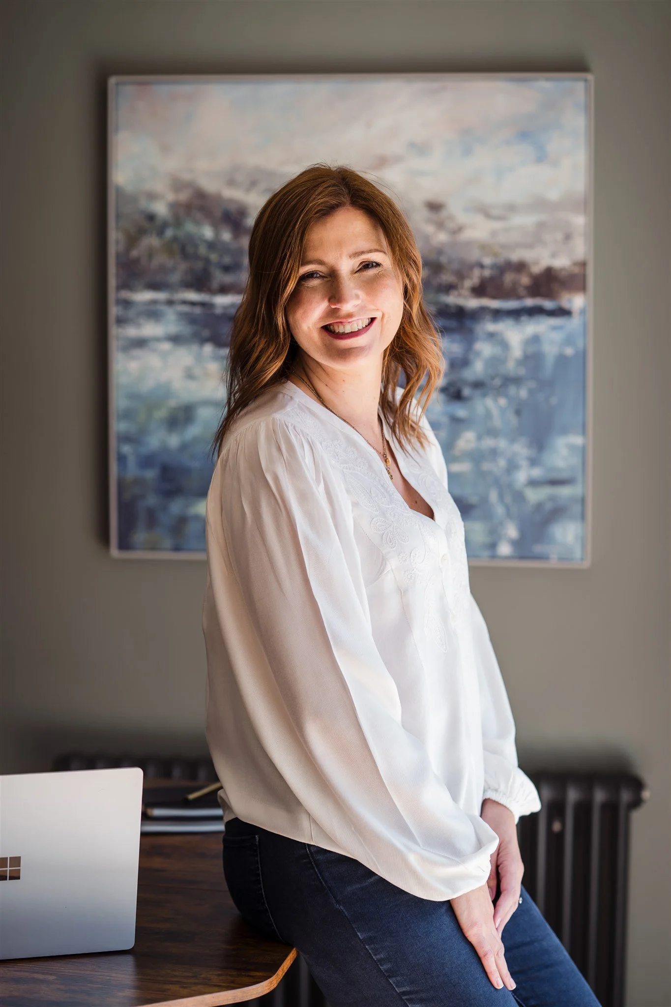 A female leadership and neurodiversity coach with shoulder-length brown hair wearing a white blouse and blue jeans, smiling while sitting on a wooden table in a room with a painting in the background.
