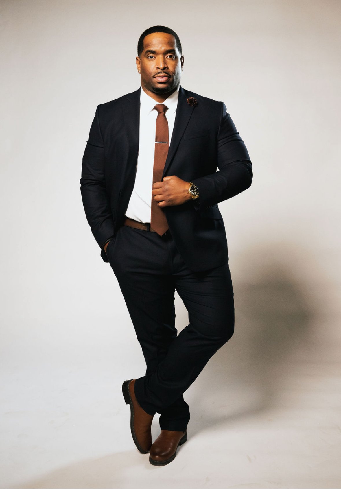 A man in a black suit, white shirt, and brown tie, standing with one hand in his pocket and the other adjusting his jacket, against a neutral background.