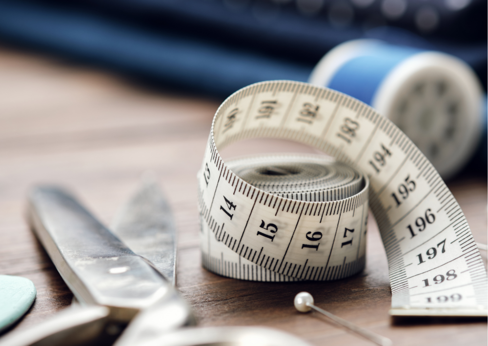 Close-up of tailoring tools including a rolled measuring tape, scissors, a sewing pin, and thread spool.