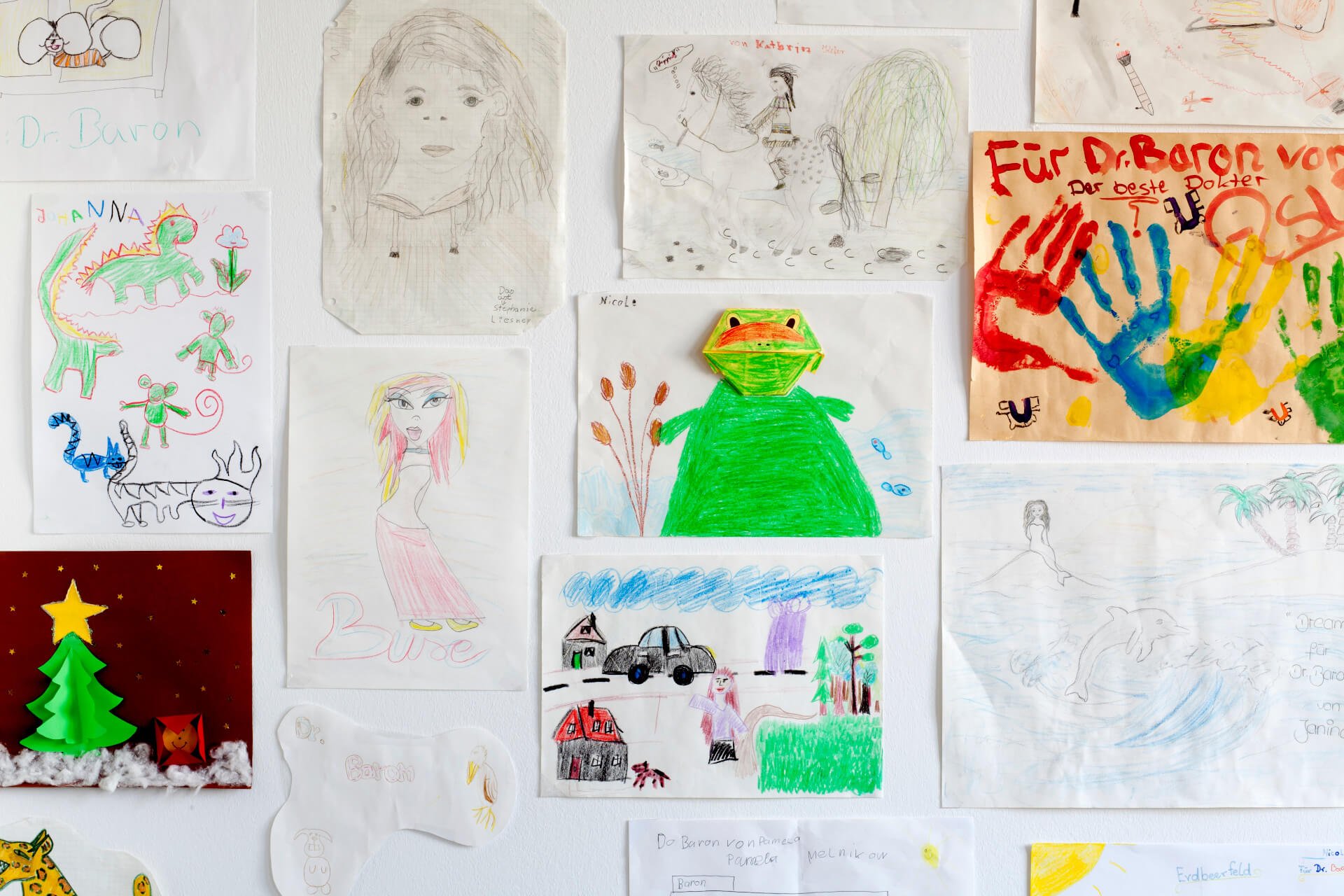 Verschiedene Kinderzeichnungen und Gemälde, darunter eine farbige Zeichnung eines Frosches, eine Silhouette einer Frau, ein bunter Dinosaurier sowie einen Weihnachtsbaum, alles an einer weißen Wand befestigt.