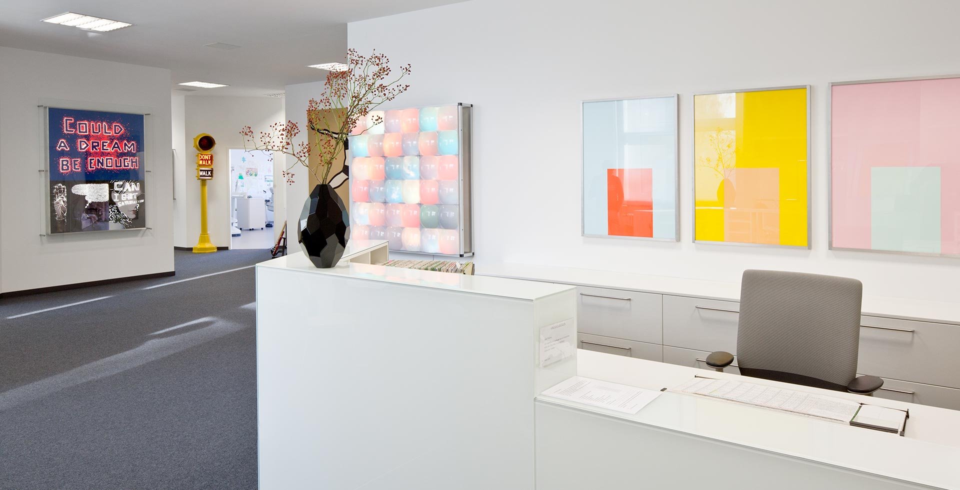 Heller Büroempfang mit weißen Möbeln, Wandkunst, Vase mit Zweigen, grauem Stuhl und Farbfeldern an der Wand.