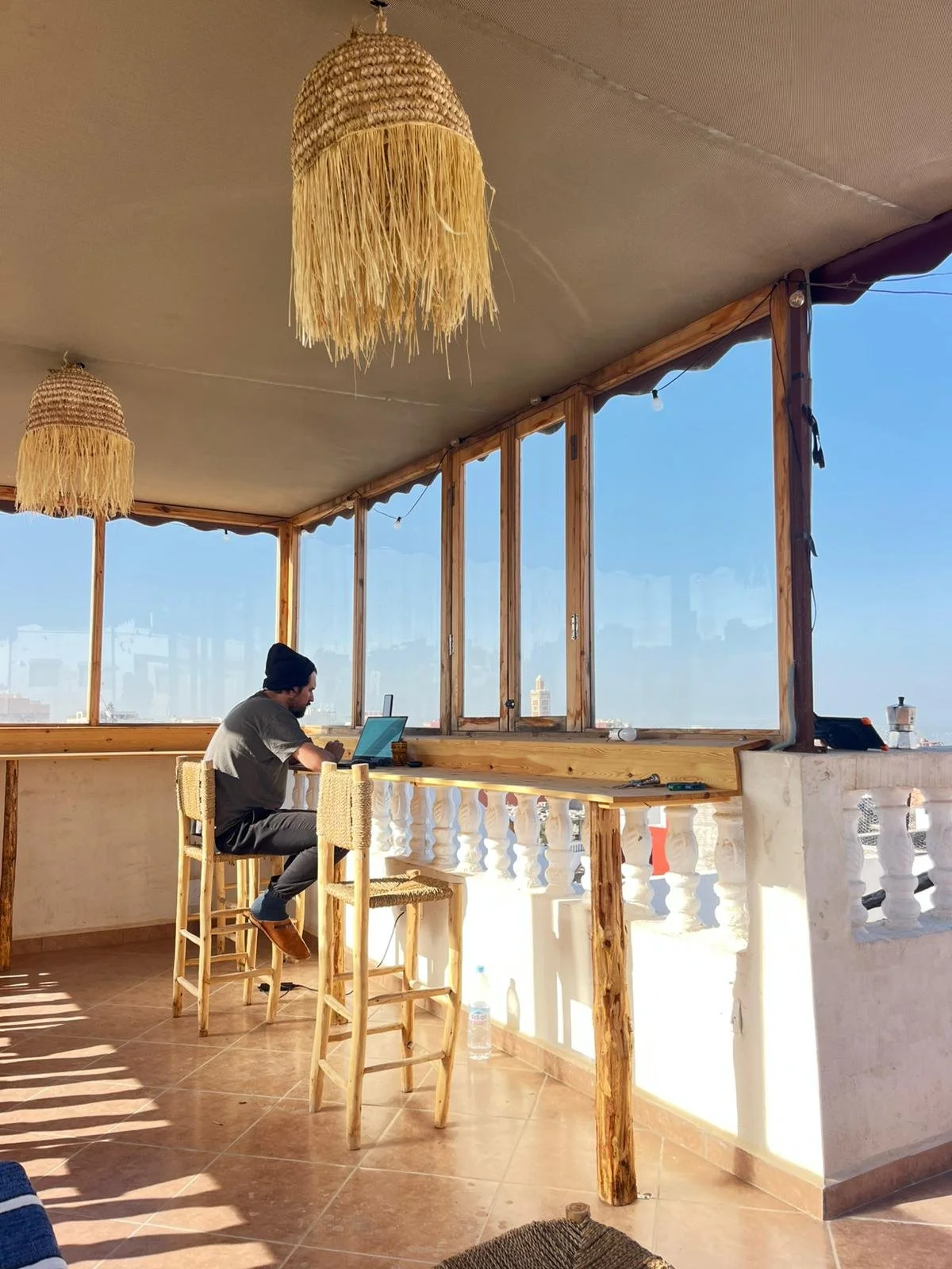 A man sitting on a barstool working on a laptop in a bright, open space with large windows and a city view. The room has wooden accents and hanging woven lamps.