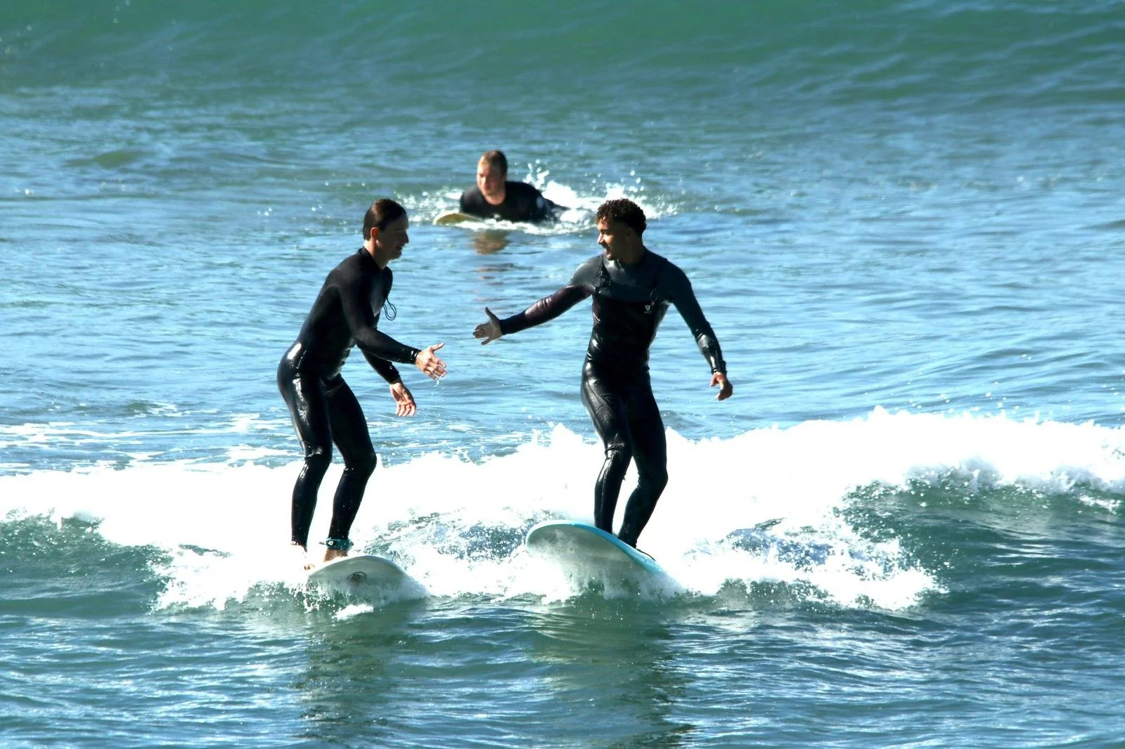 Two young men in wetsuits surfing on a wave, one giving a high five to the other, with a person in the background swimming in the ocean.
