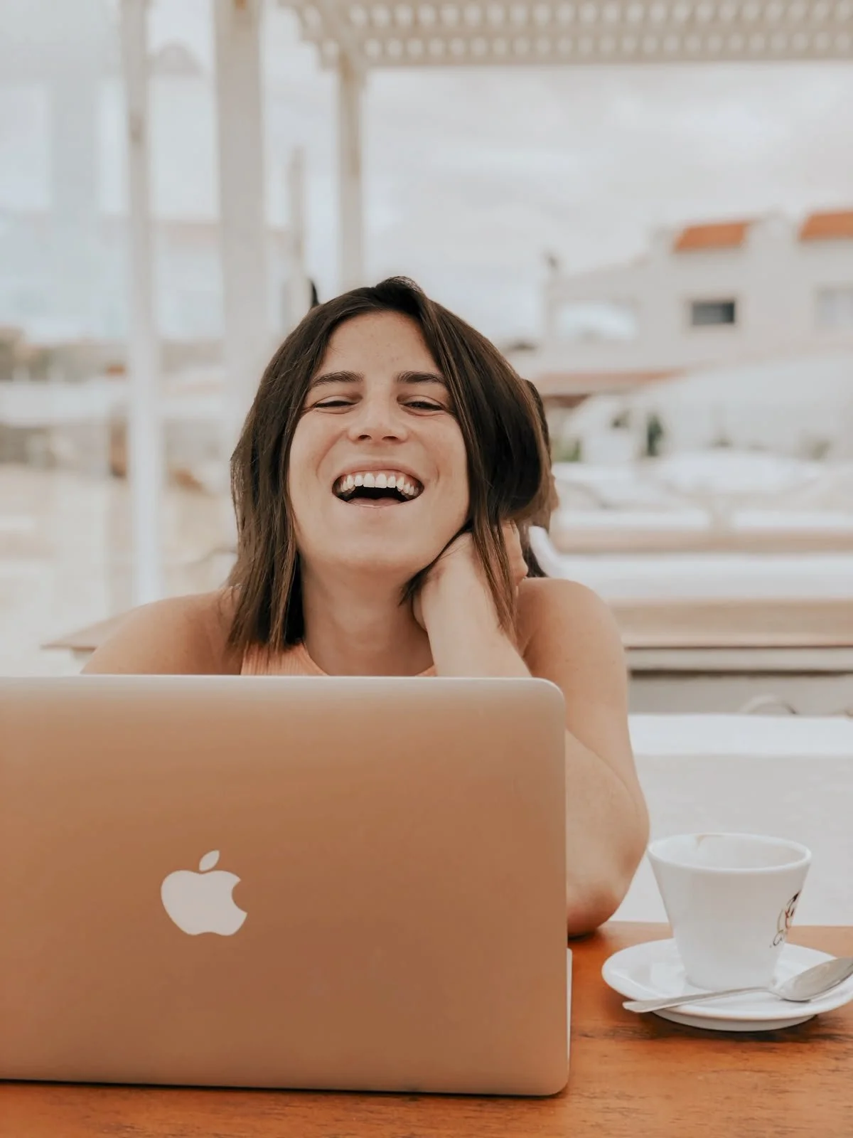 A woman with short brown hair laughing in a cafe, sitting at a wooden table with a silver MacBook, a white coffee cup, and a saucer.