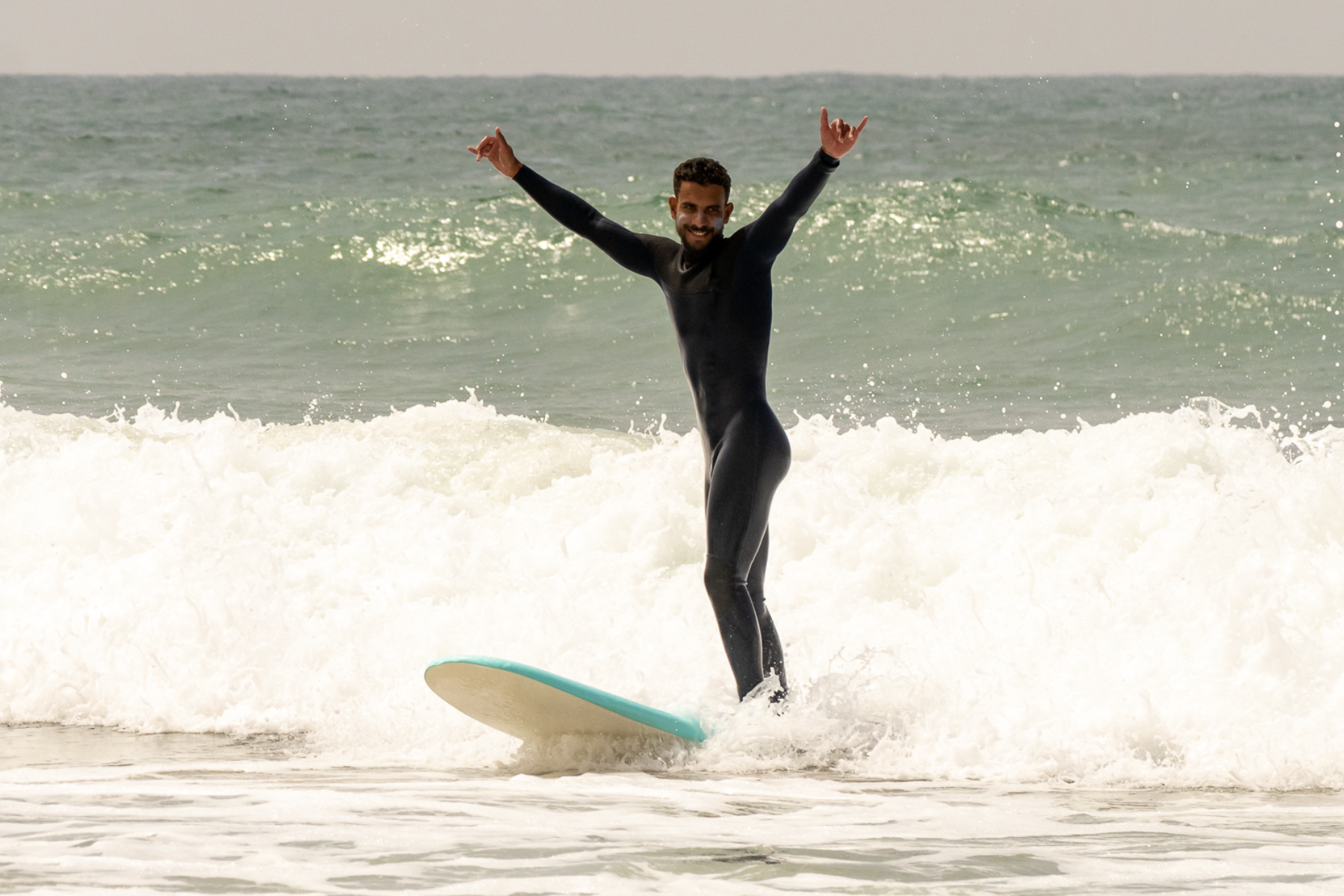 Man in a black wetsuit riding a surfboard on a wave at the beach, smiling with arms raised.