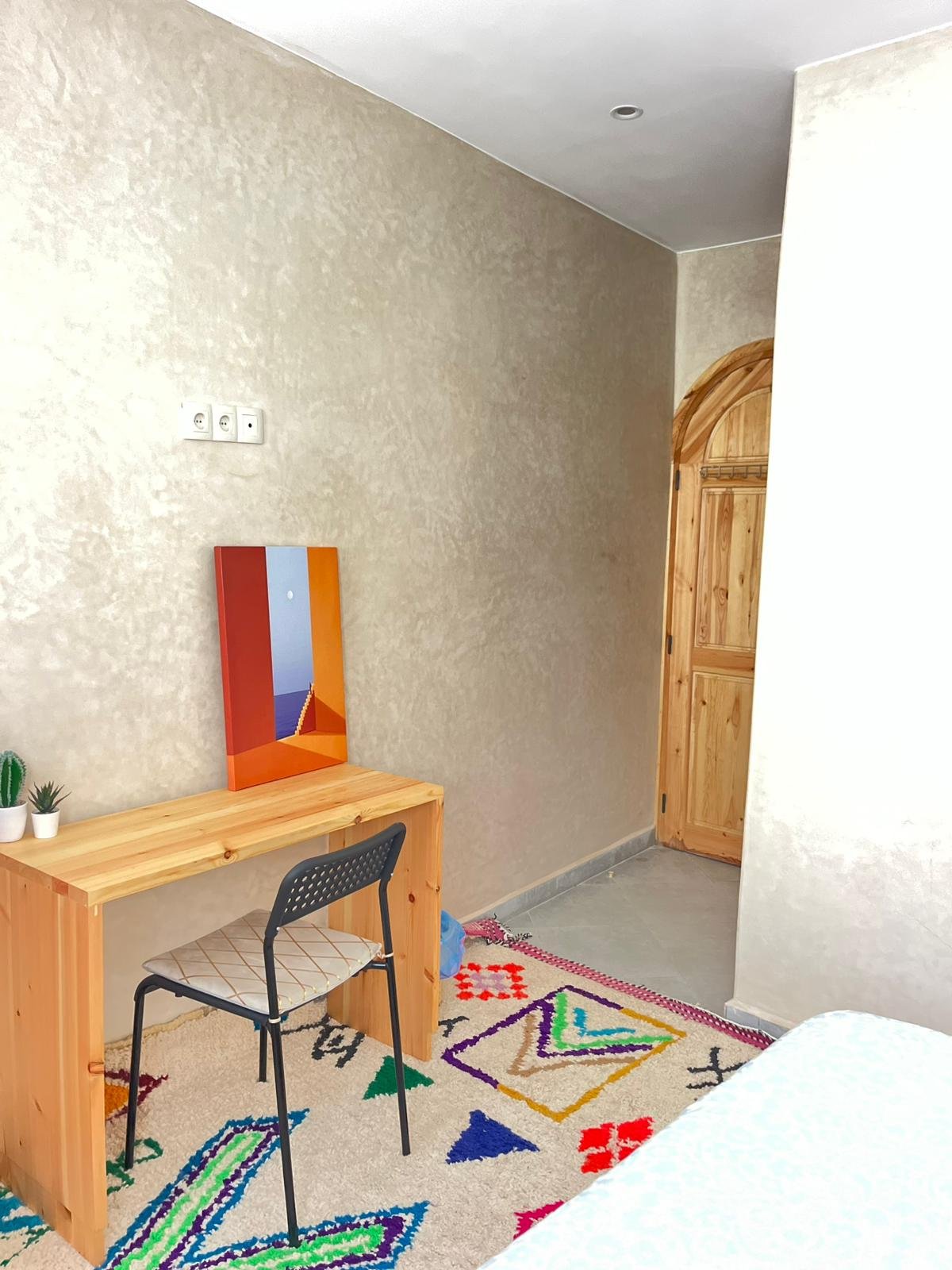 Interior of a room with beige walls, a wooden door, a wooden desk with a mirror, a black chair with a patterned cushion, a colorful rug, and some small potted plants.