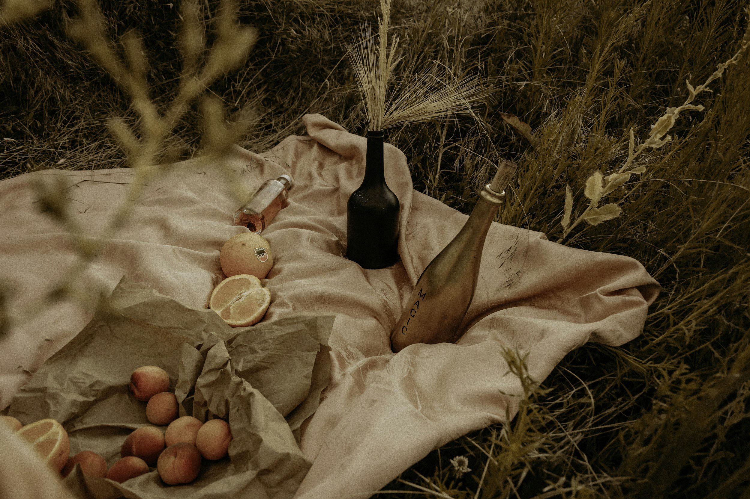 A picnic setup in a field with a pink satin cloth, featuring a black vase with pampas grass, a champagne bottle, a small bottle of perfume or oil, peaches, a sliced orange, and a crumpled paper bag with more peaches inside.