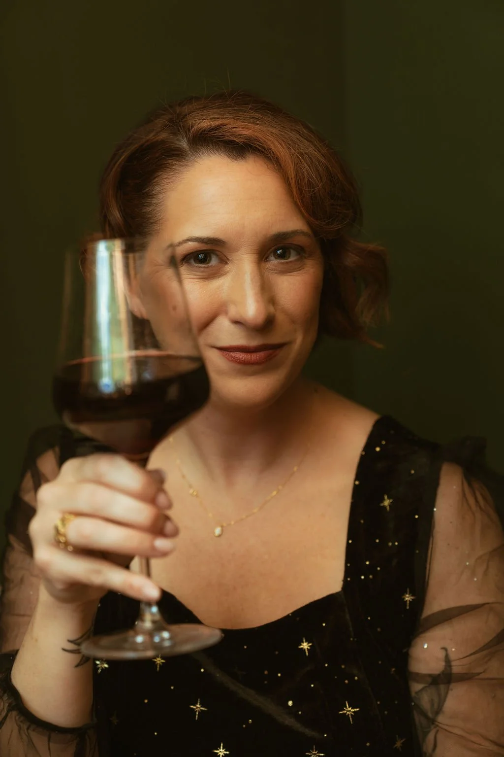 A woman with short red hair holding a glass of red wine, smiling at the camera, wearing a black dress with gold star patterns and a gold necklace.