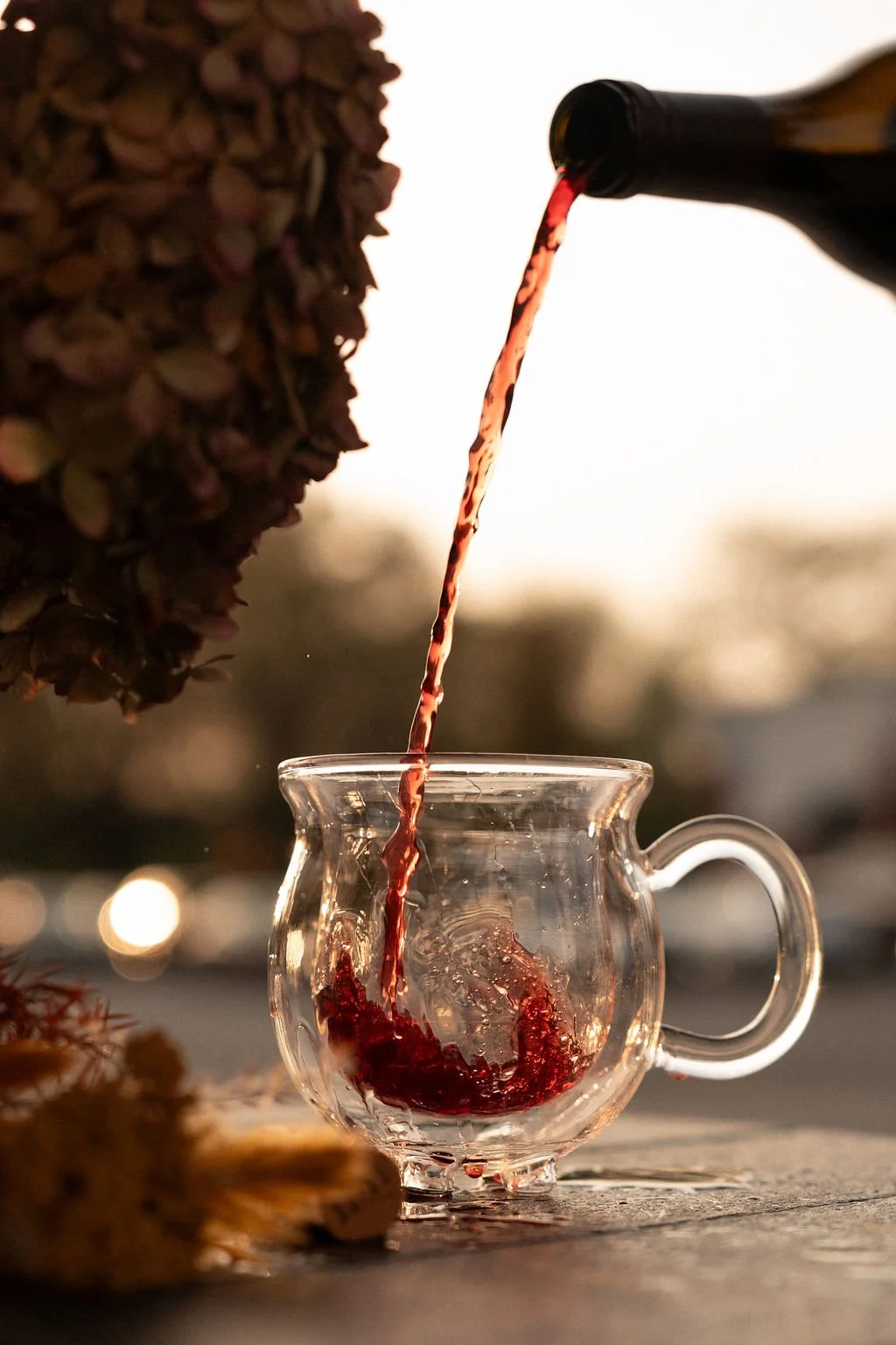 Red wine being poured into a clear glass cup on an outdoor surface with blurred background and sunset lighting.
