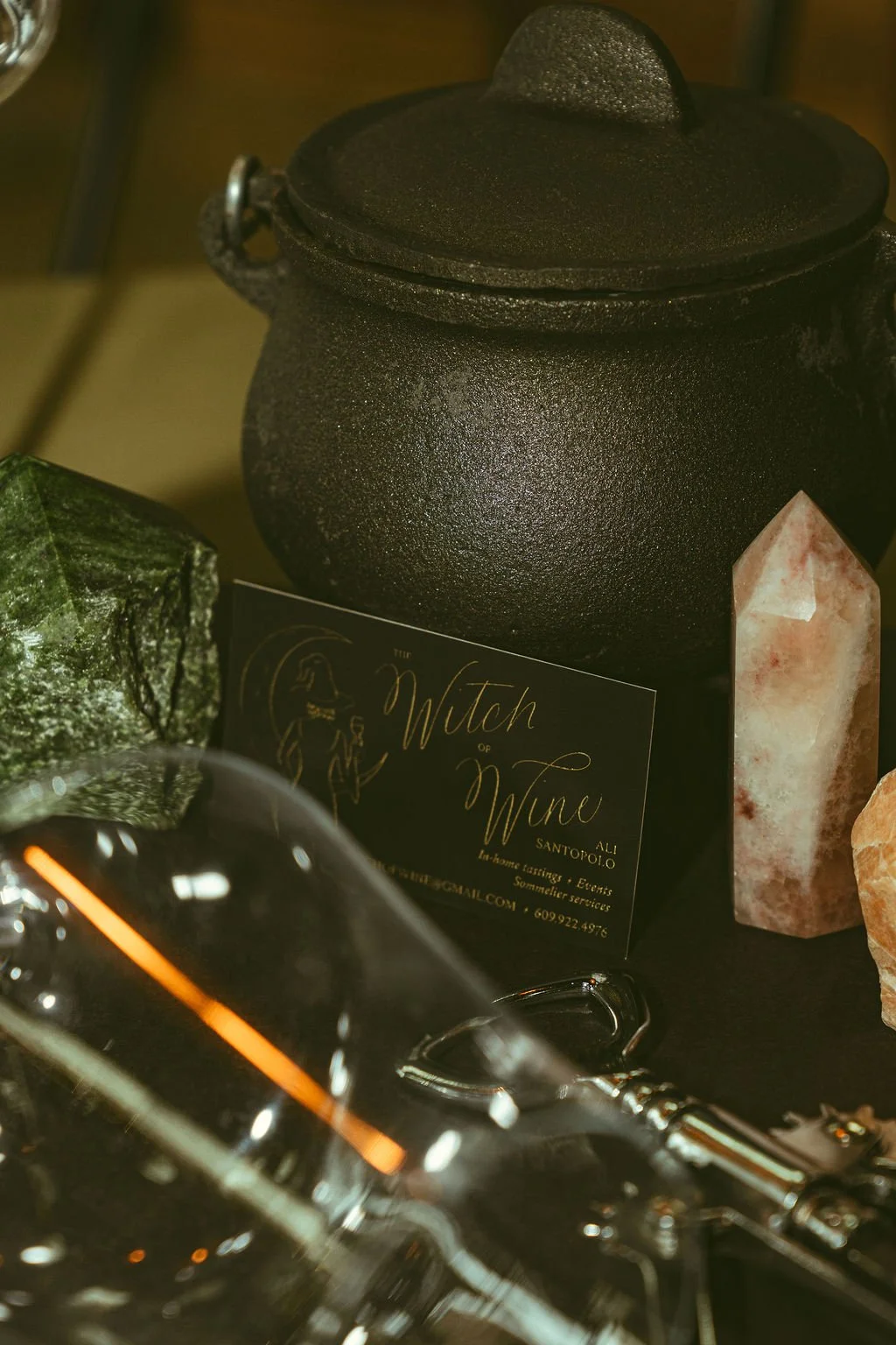 A black cast iron pot with a lid, a business card titled 'The Witch of Wine' by Ali Santopolo, partially visible green stone, and a large pinkish-orange crystal.